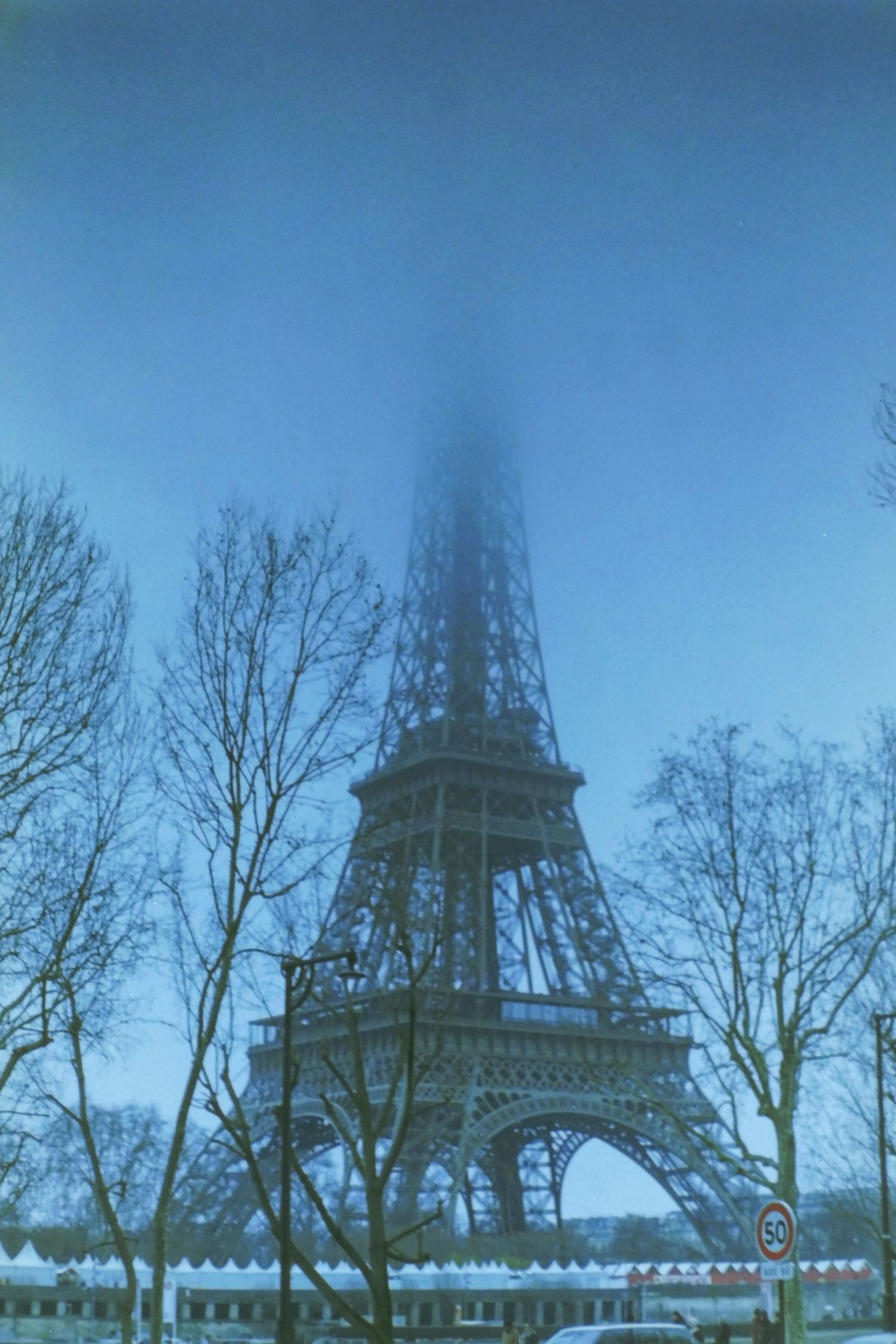 The Eiffel Tower in Paris emerges through dense fog surrounded by bare winter trees on a blue misty day.