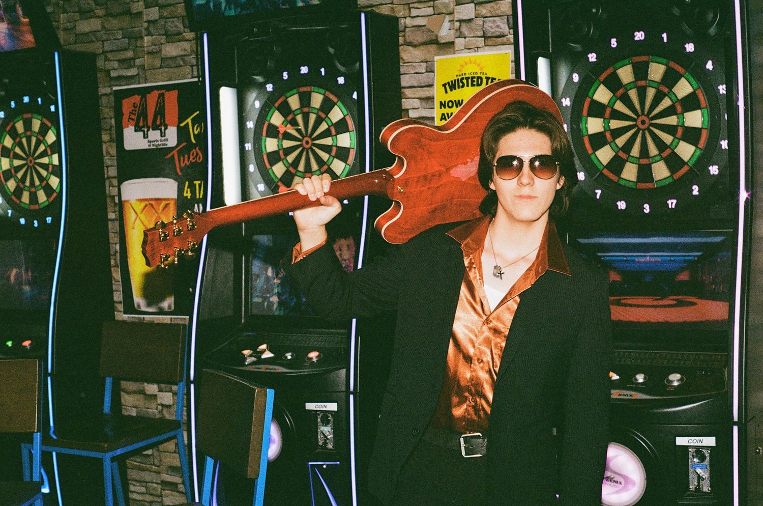 Rockstar posing with electric guitar on shoulder wearing sunglasses in a bar with dartboards.
