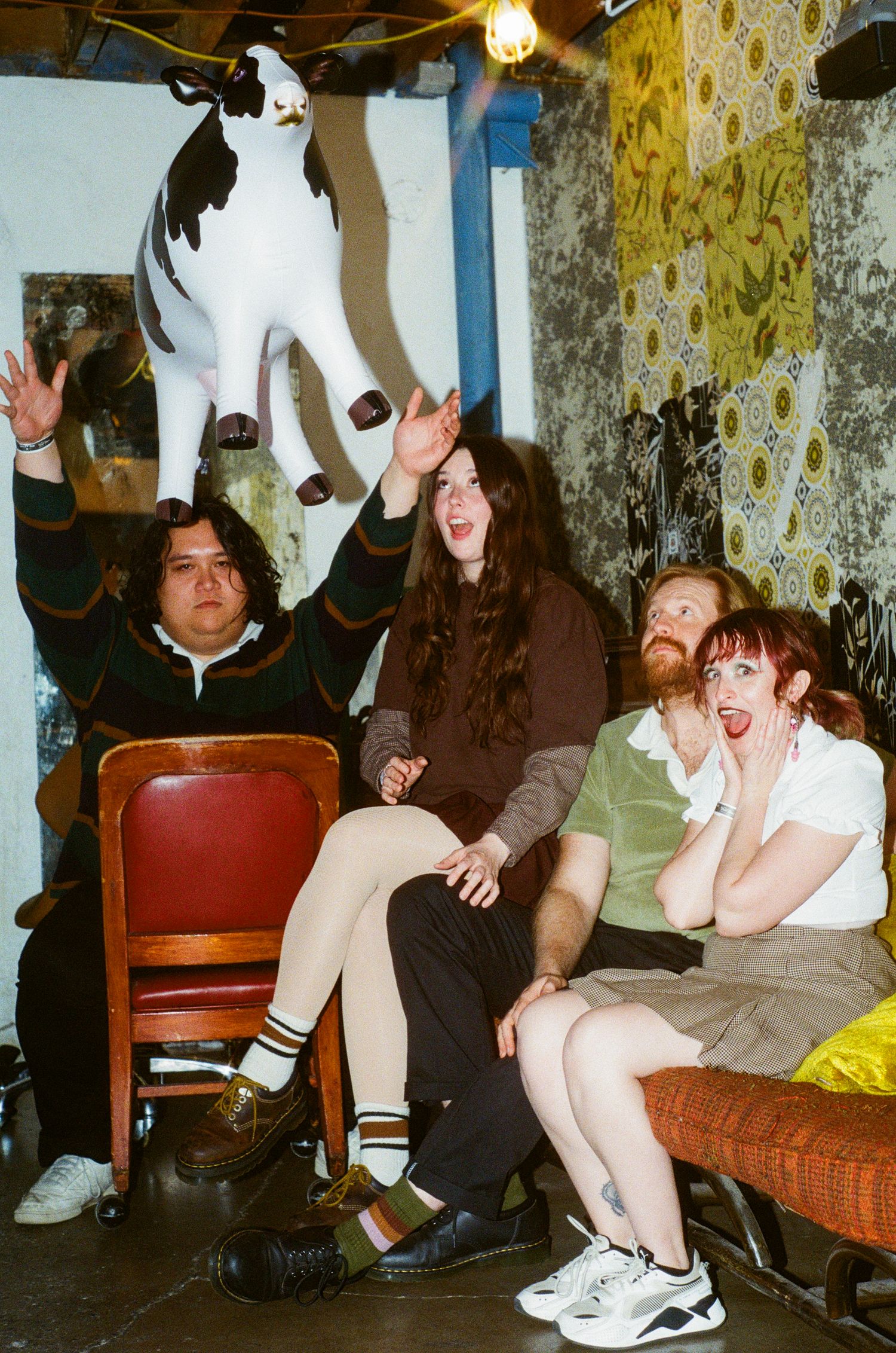 Four friends laughing and tossing inflatable cow in eclectic decorated room.