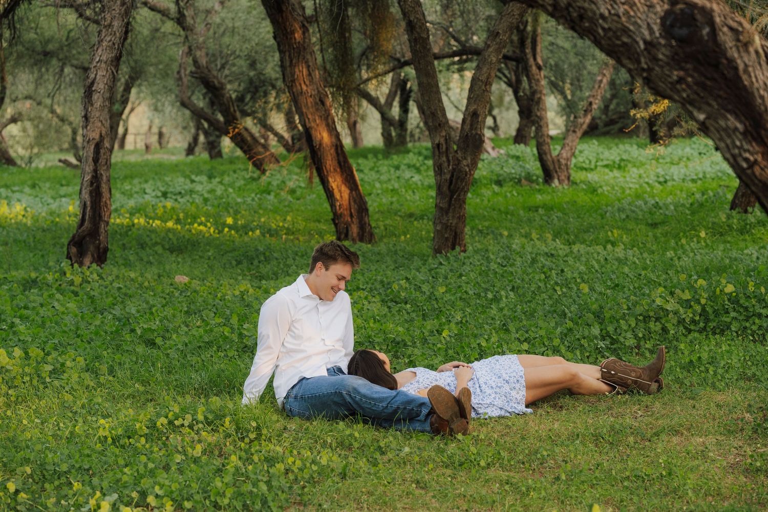 Couple relaxing together on lush green grass surrounded by trees in a peaceful outdoor park setting.
