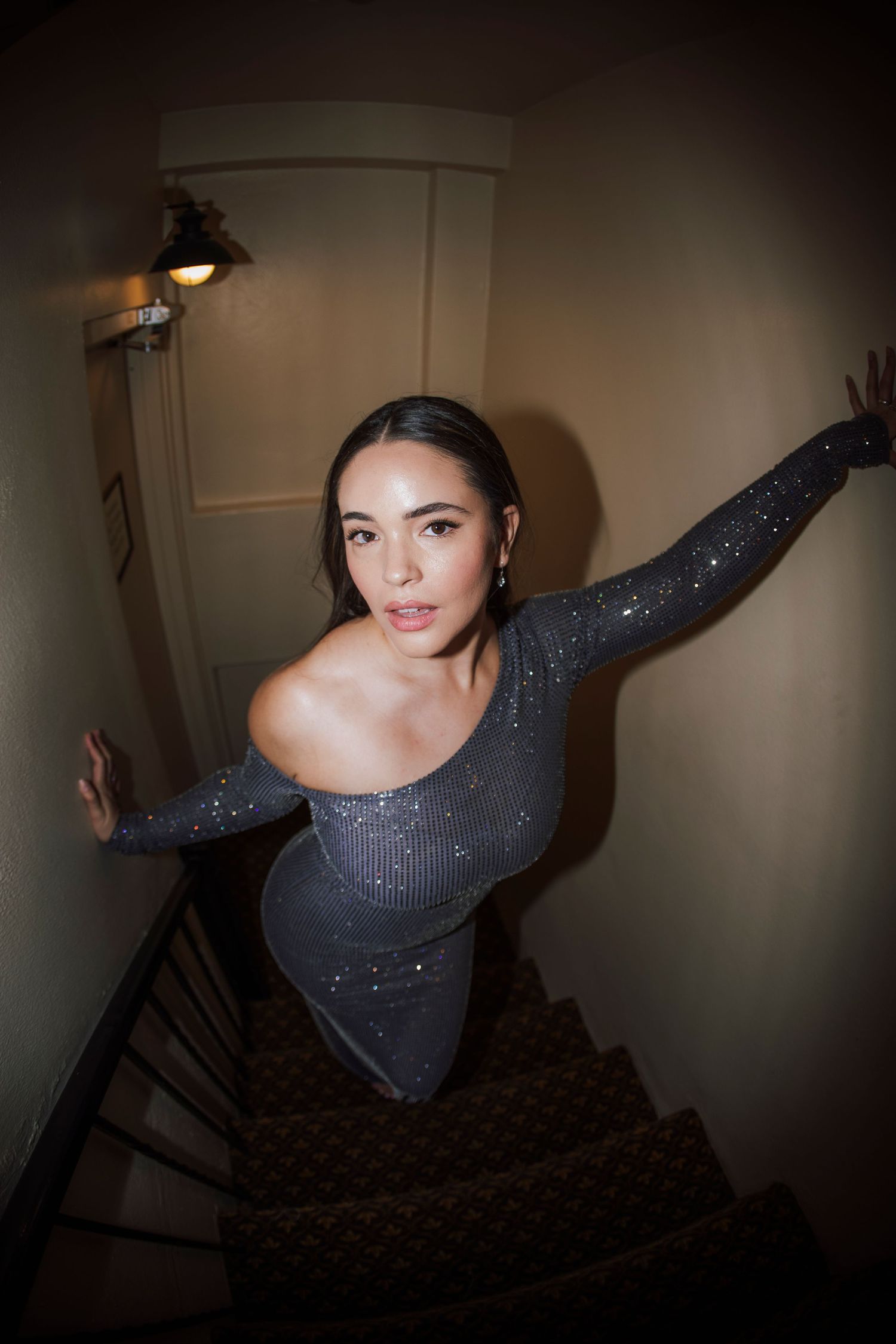 Woman in sparkly gray off-shoulder dress poses on dimly lit staircase.