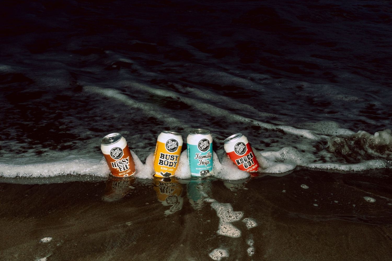 Four colorful beer cans lined up on a dark sandy beach with ocean waves washing around them at night.