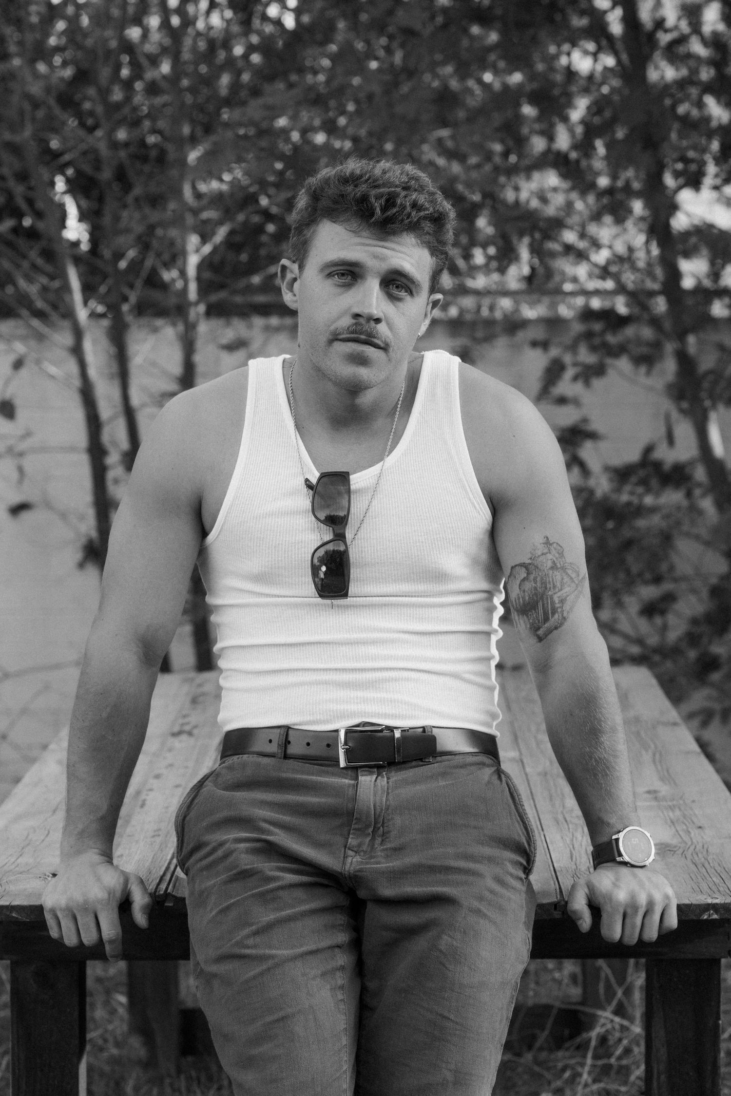Black and white portrait of muscular man with mustache in white tank top seated outdoors.