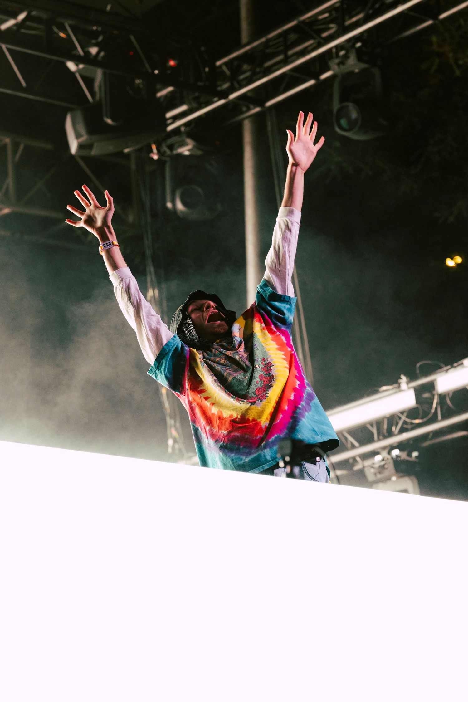 Energetic performer in colorful tie-dye shirt raising both arms on stage with backlit fog.