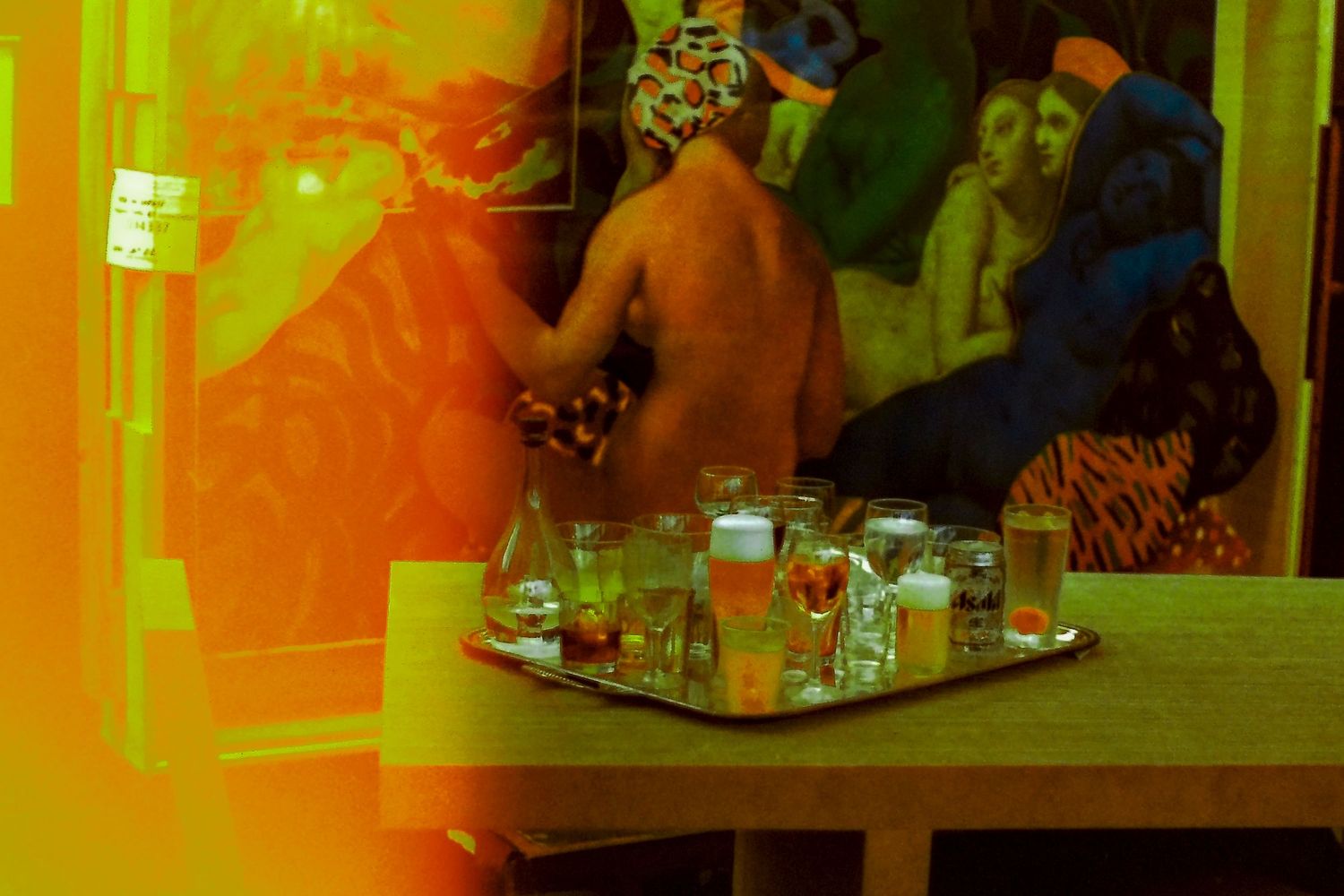 Person in colorful mask near a tray of bottles and glassware on a wooden table in a warmly lit room.