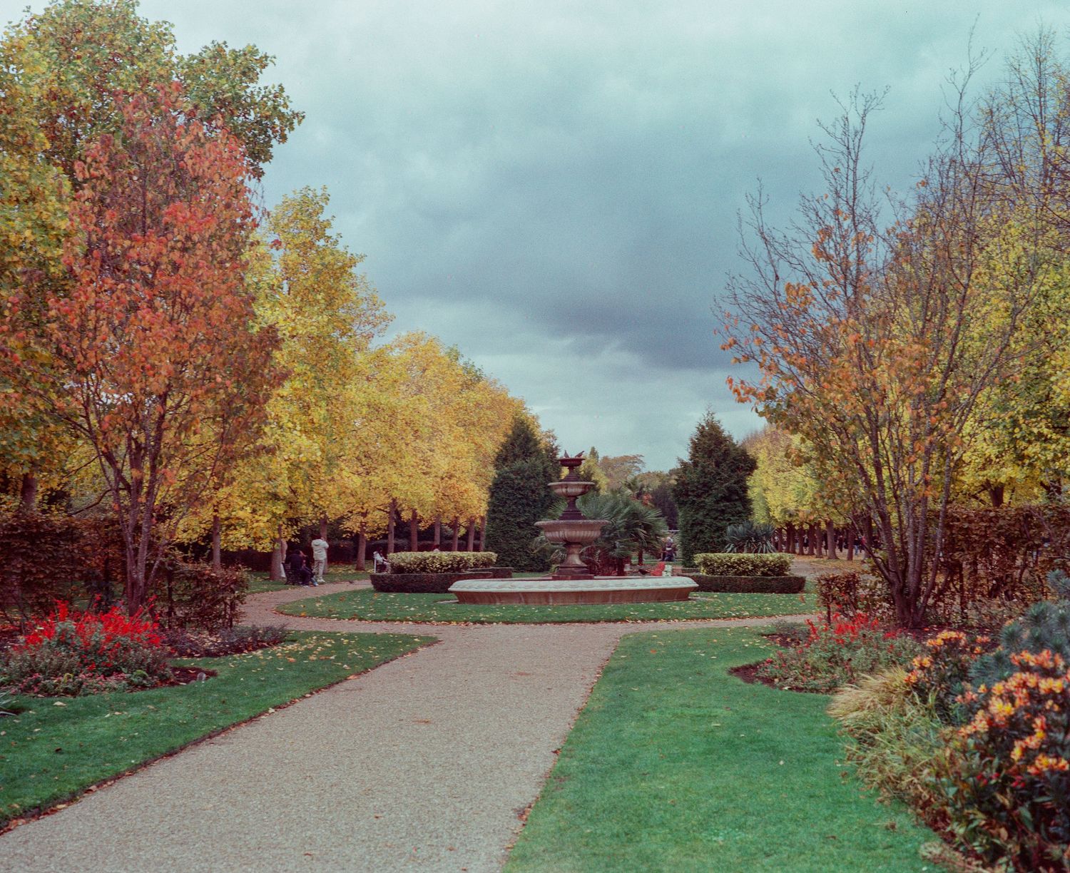 Autumn park pathway leading to ornate fountain surrounded by colorful fall foliage trees under cloudy sky.