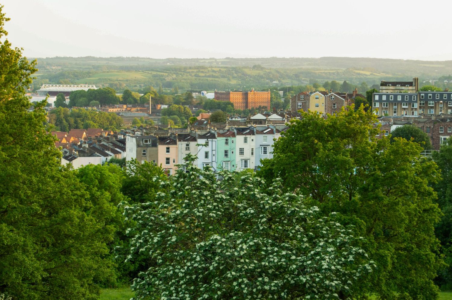 Aerial view of colorful townhouses and rooftops nestled among lush green trees with rolling hills beyond.