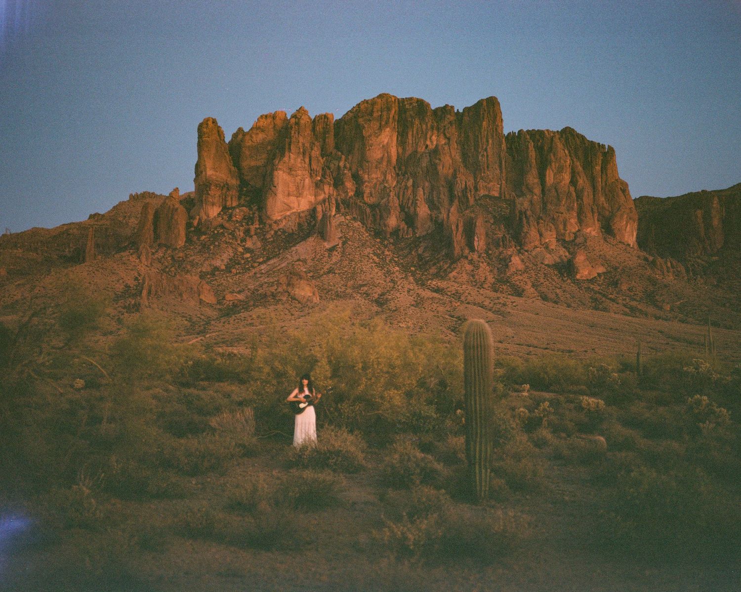 Woman playing guitar in Arizona desert at dusk with Superstition Mountains and saguaro cactus backdrop.