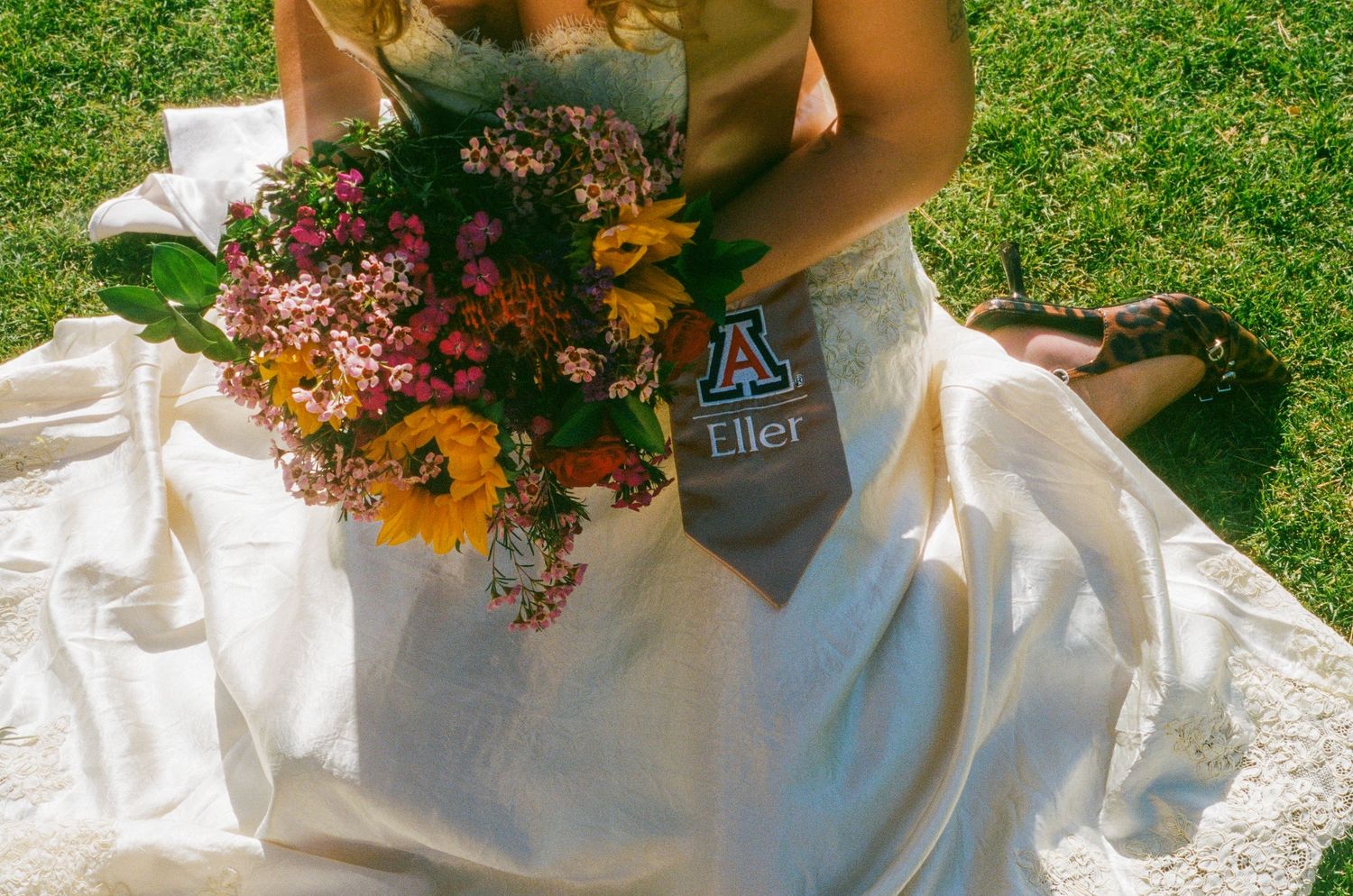 Bride in white gown sitting on grass holding colorful wildflower bouquet with University of Arizona Eller sash.