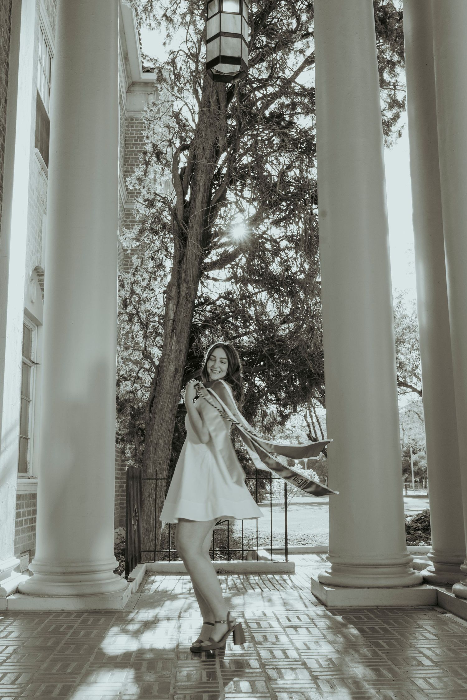 Woman in white dress posing between tall white columns with scarf flowing in black and white photo.