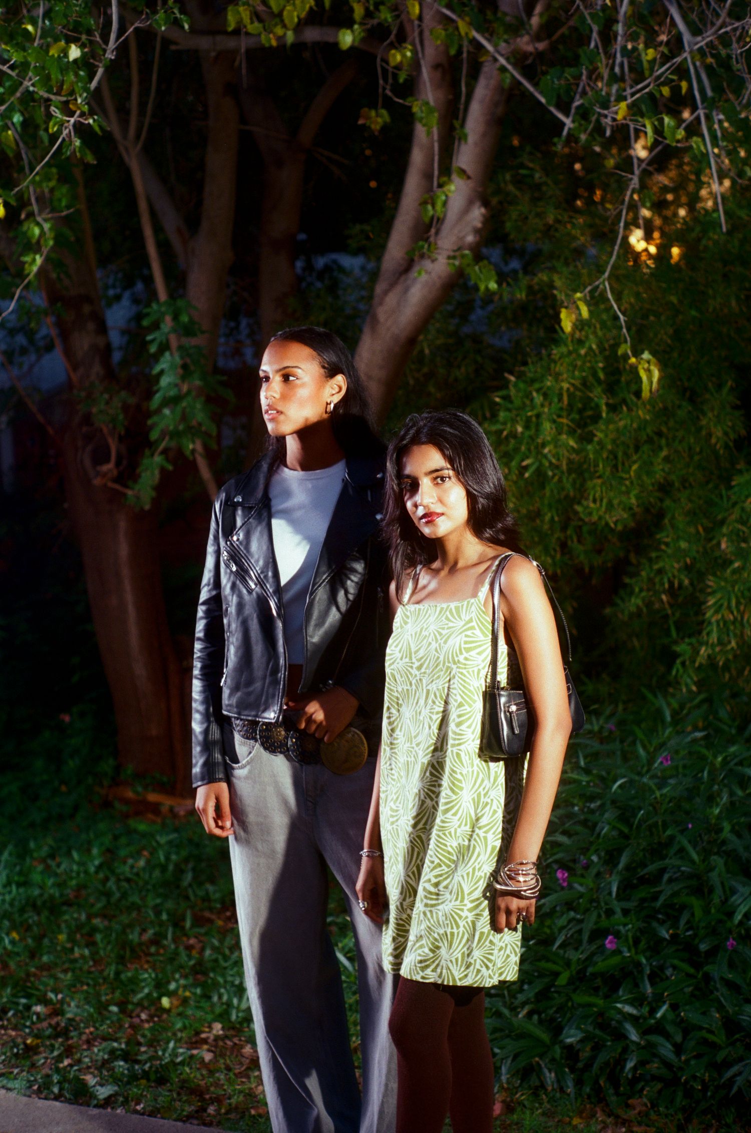 Two young women in stylish outfits pose together in a garden at night.