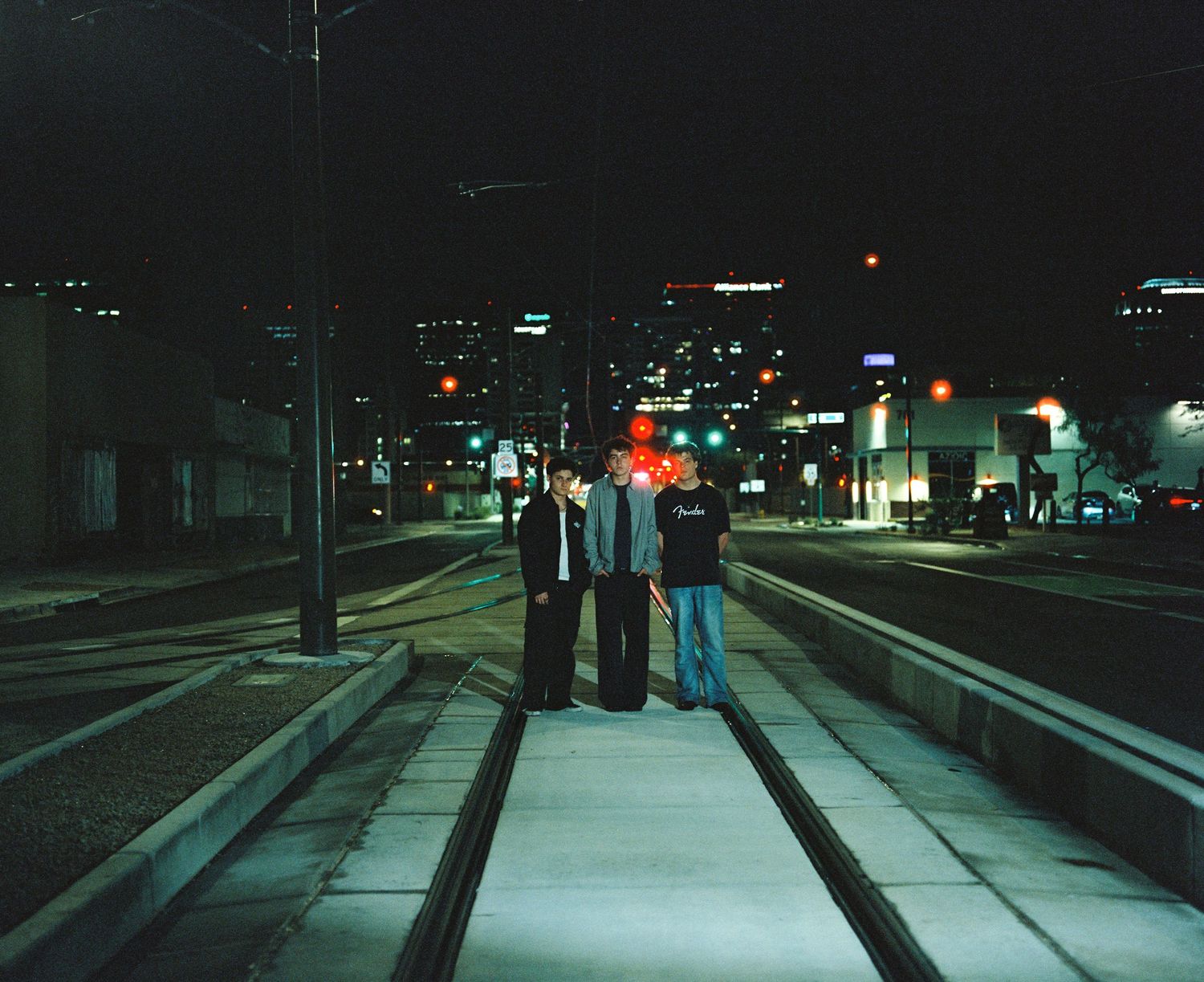 Three people standing on city train tracks at night with urban skyline behind them.