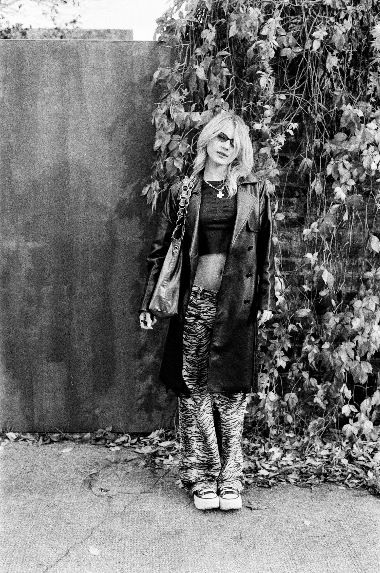 Black and white photo of blonde woman in leather coat and tiger print pants by ivy wall.