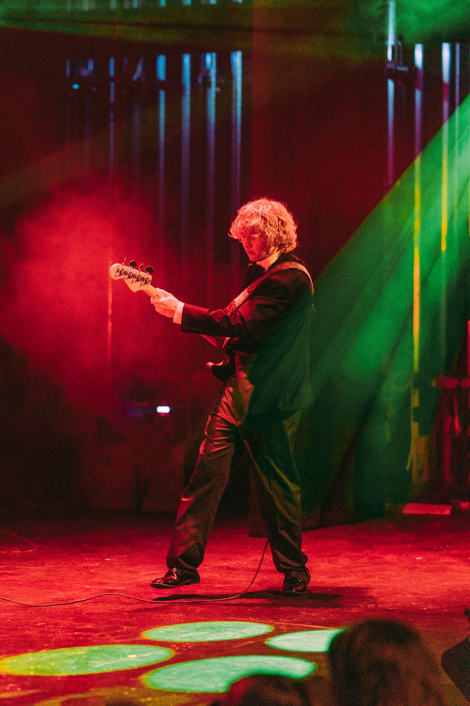 Guitarist in a black suit performing on a colorful stage with red and green lighting effects.