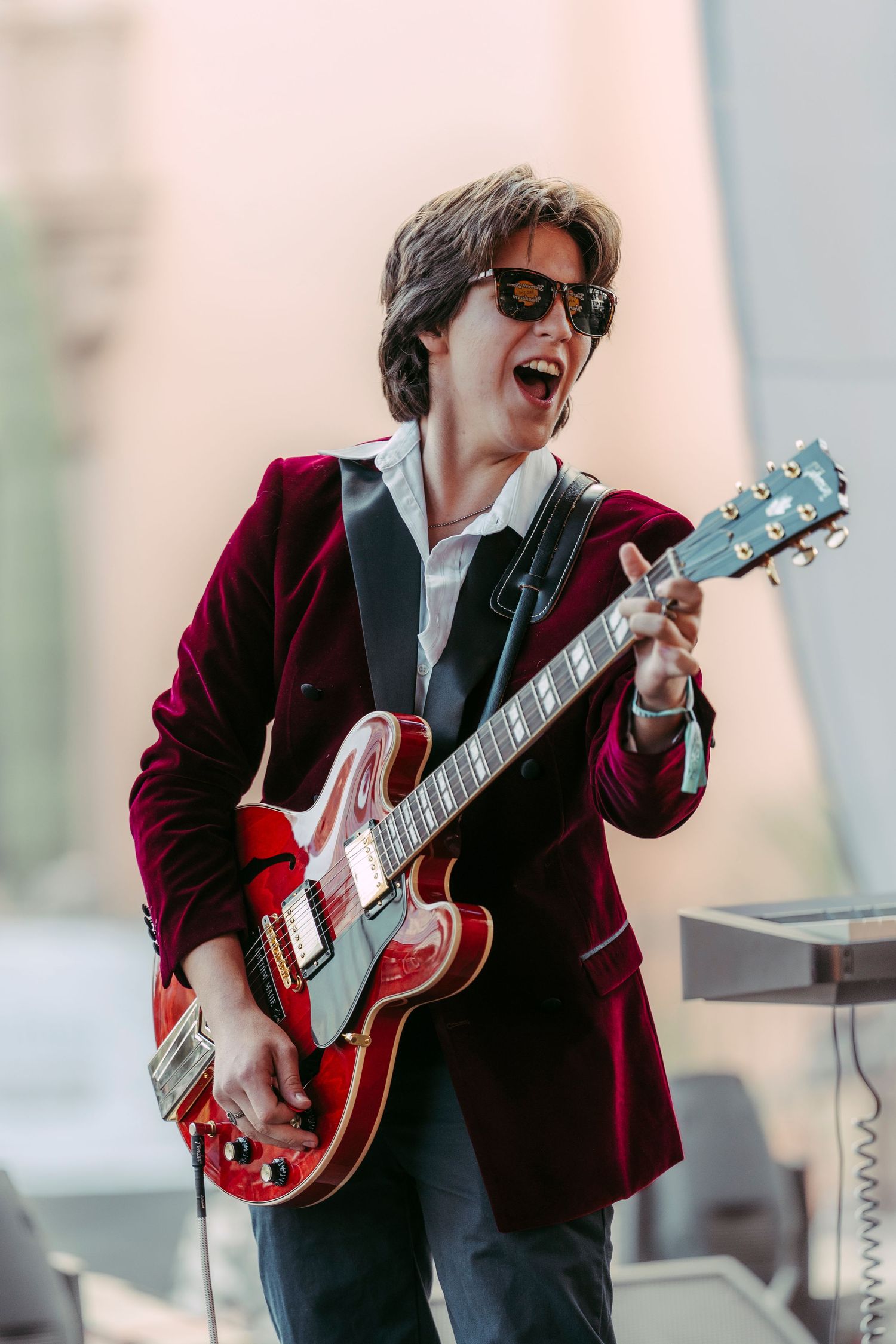 Energetic guitarist in red velvet blazer playing red semi-hollow electric guitar on outdoor stage.