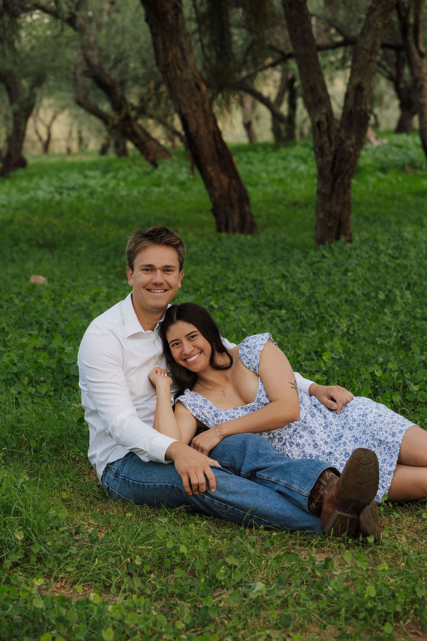 Happy couple sitting together on green grass in wooded park, smiling at camera.