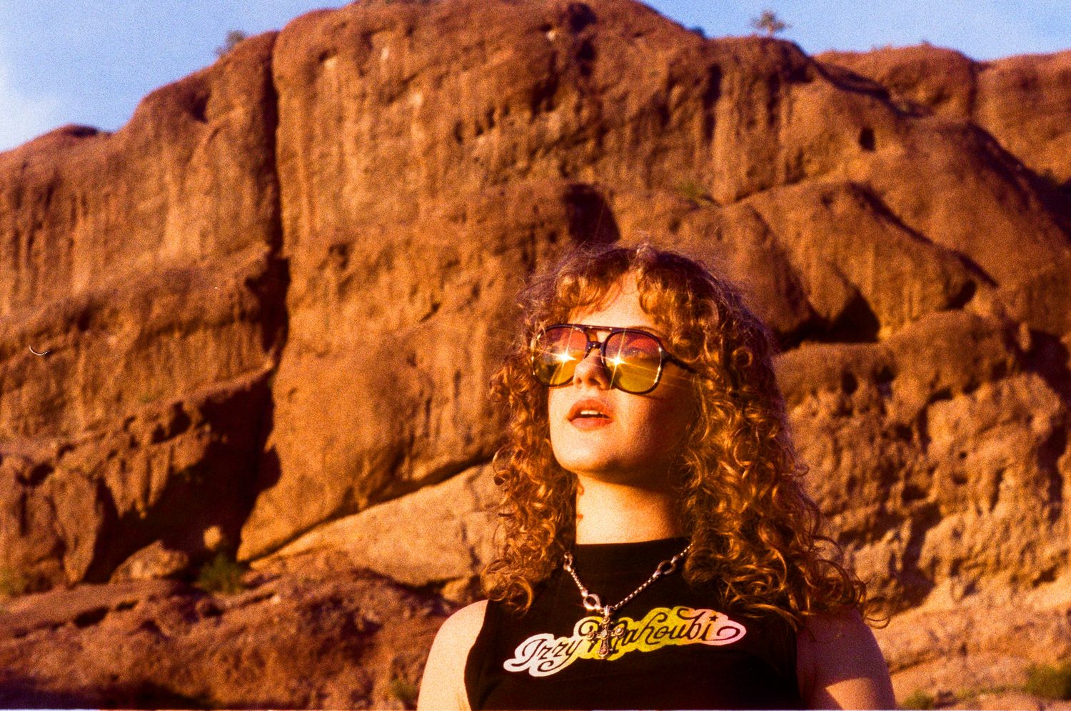 Curly-haired woman in black tank top with sunglasses poses before red rock canyon landscape.