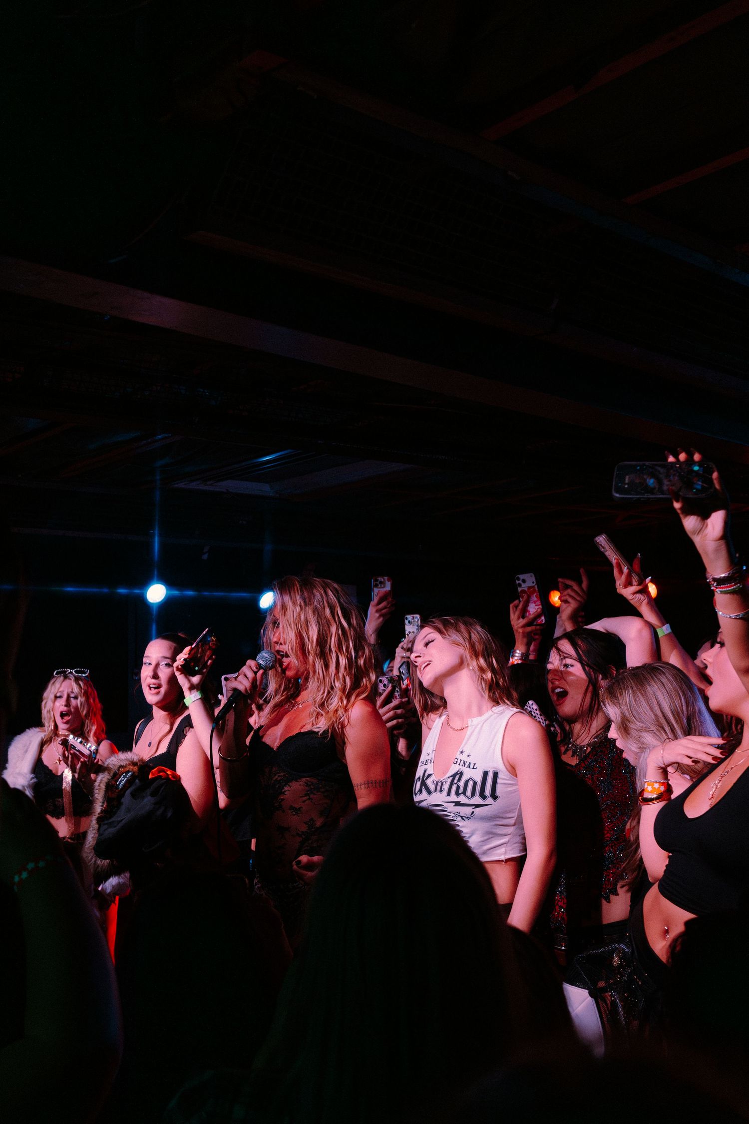 Excited crowd of women dancing and cheering at a dark concert venue, one wearing a Rock and Roll white tank top.