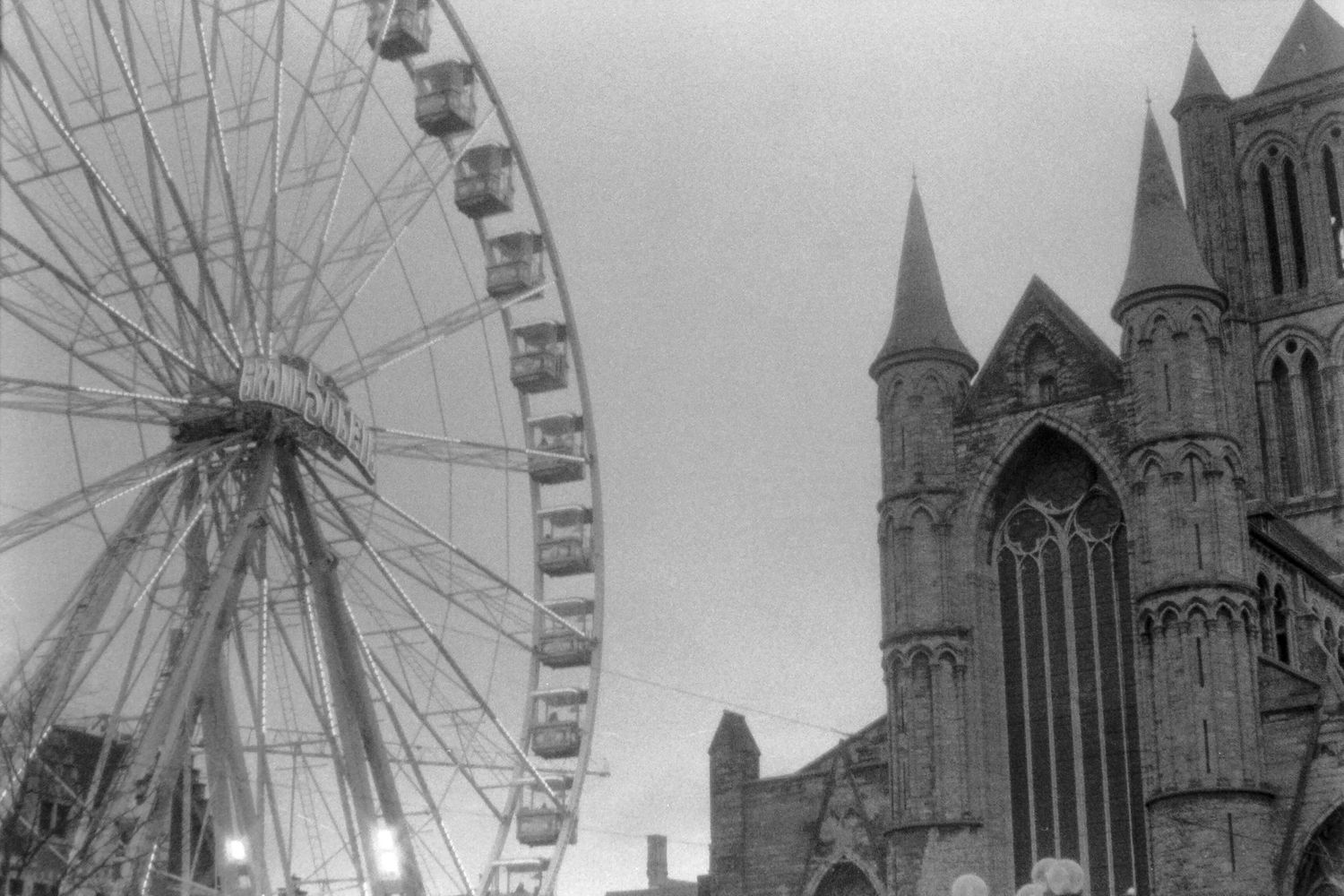 Black and white photo of a large Ferris wheel beside a Gothic church spire at a European fairground.