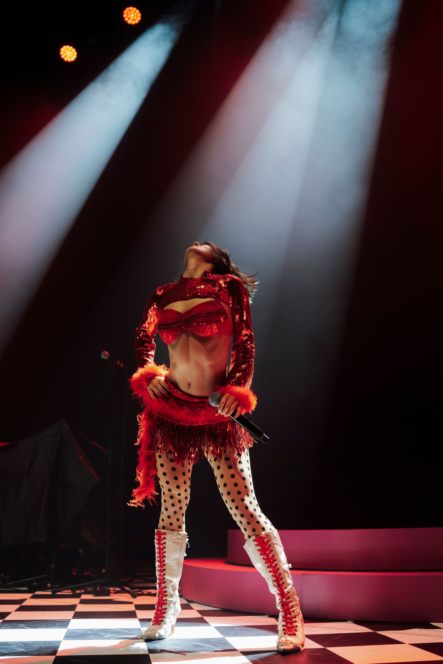 Performer in red sequined costume with polka-dot pants dances on checkered stage under dramatic spotlights.