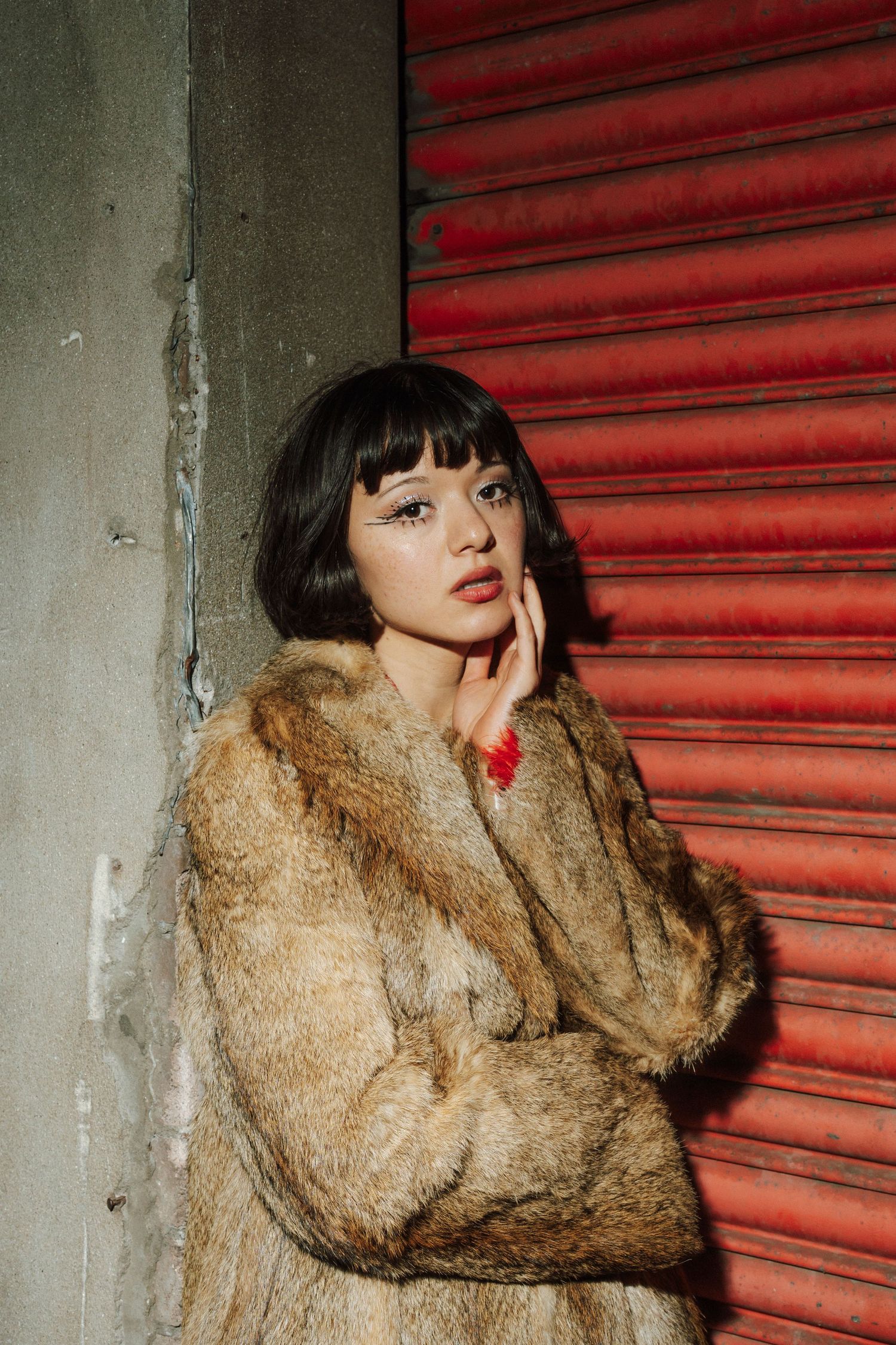 Young woman in a brown fur coat poses against a red metal shutter wall with dramatic lighting.