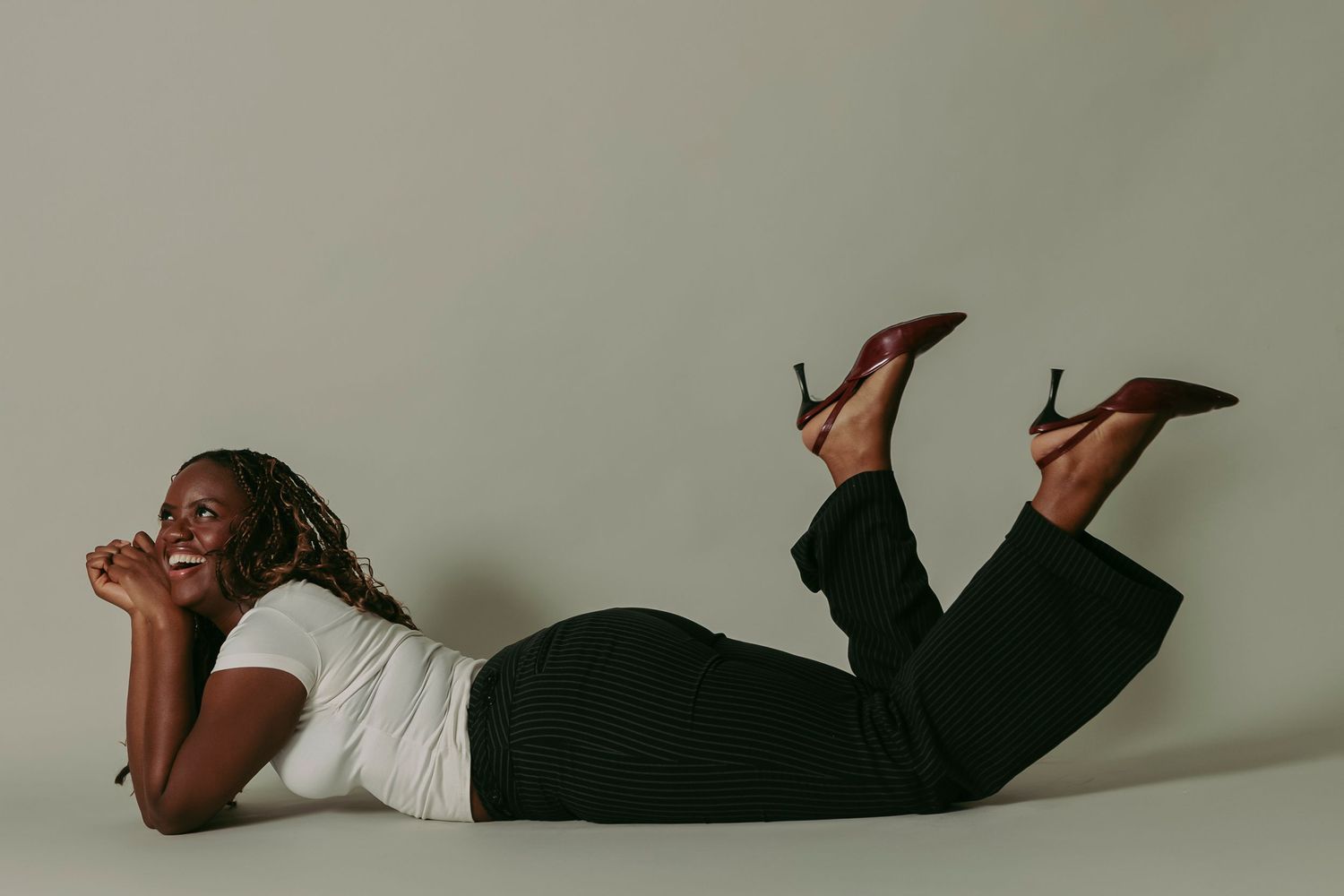 Joyful Black woman with braids lying on her stomach on a studio floor wearing black pants and heels.