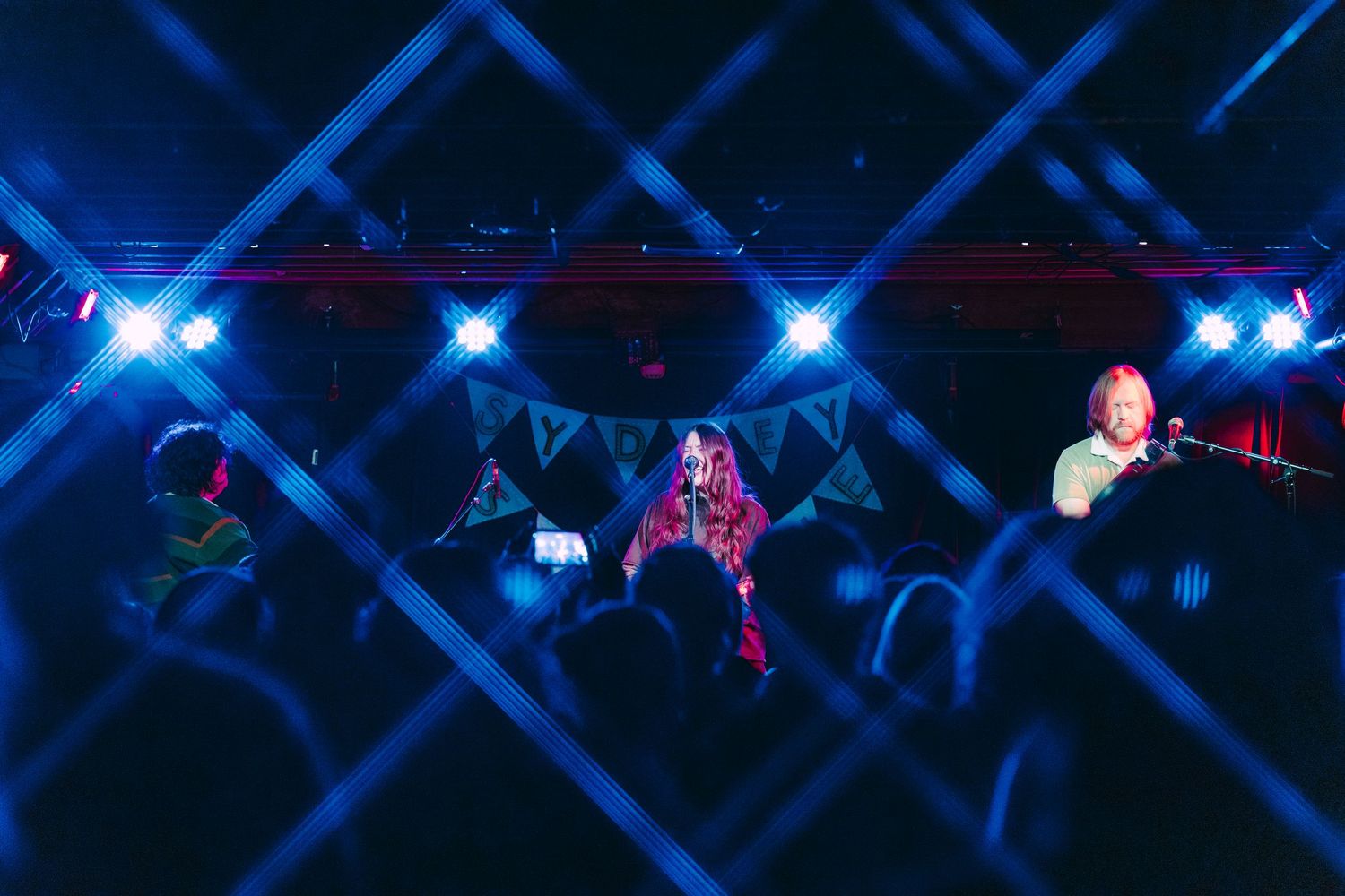 Band performing on stage with colorful blue lights and crowd gathered at indoor music venue concert.