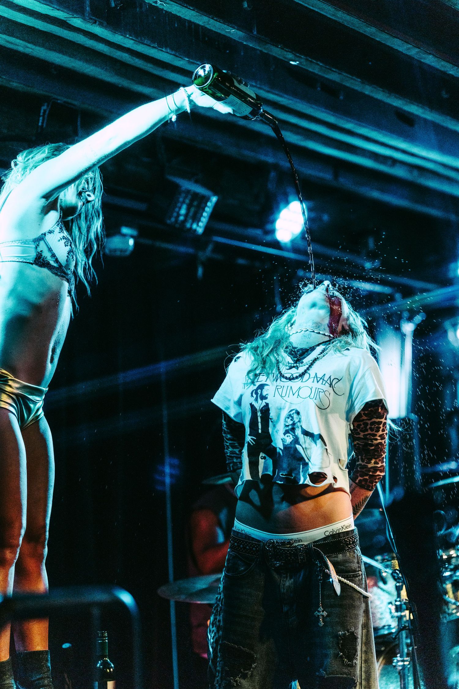Female performer pouring liquid from bottle onto another musician during wild rock concert performance on stage.