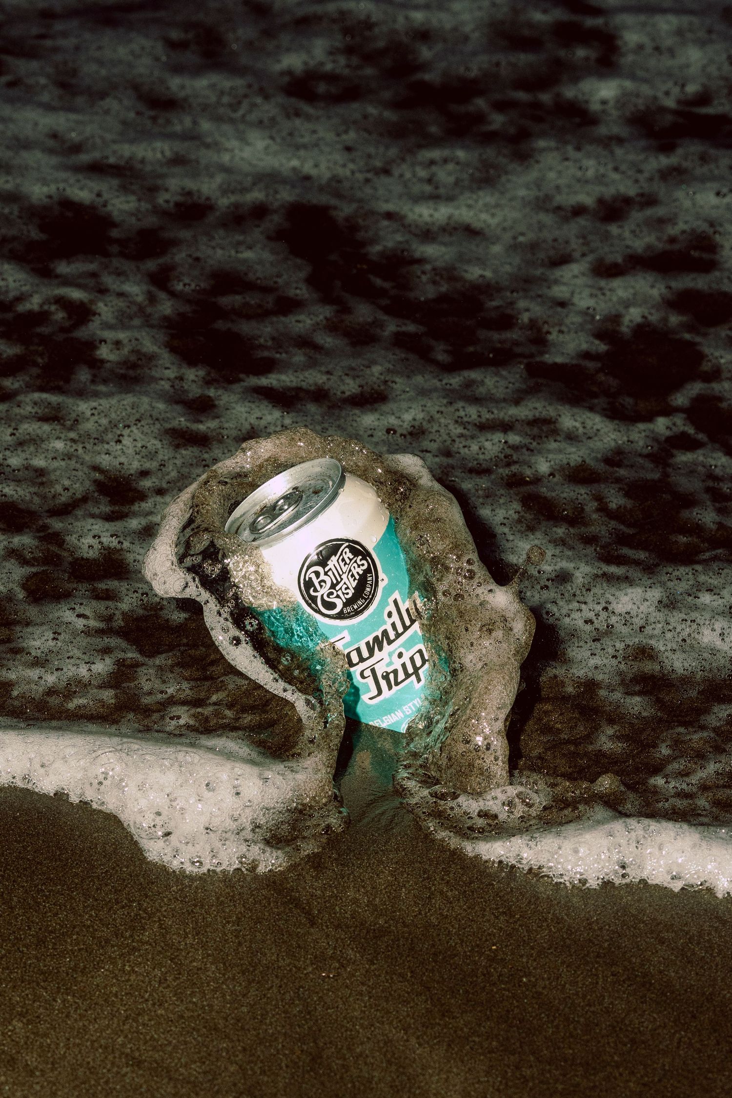 A Peculiar Brewing Family Trip beer can washed ashore on a sandy beach with ocean waves surrounding it.