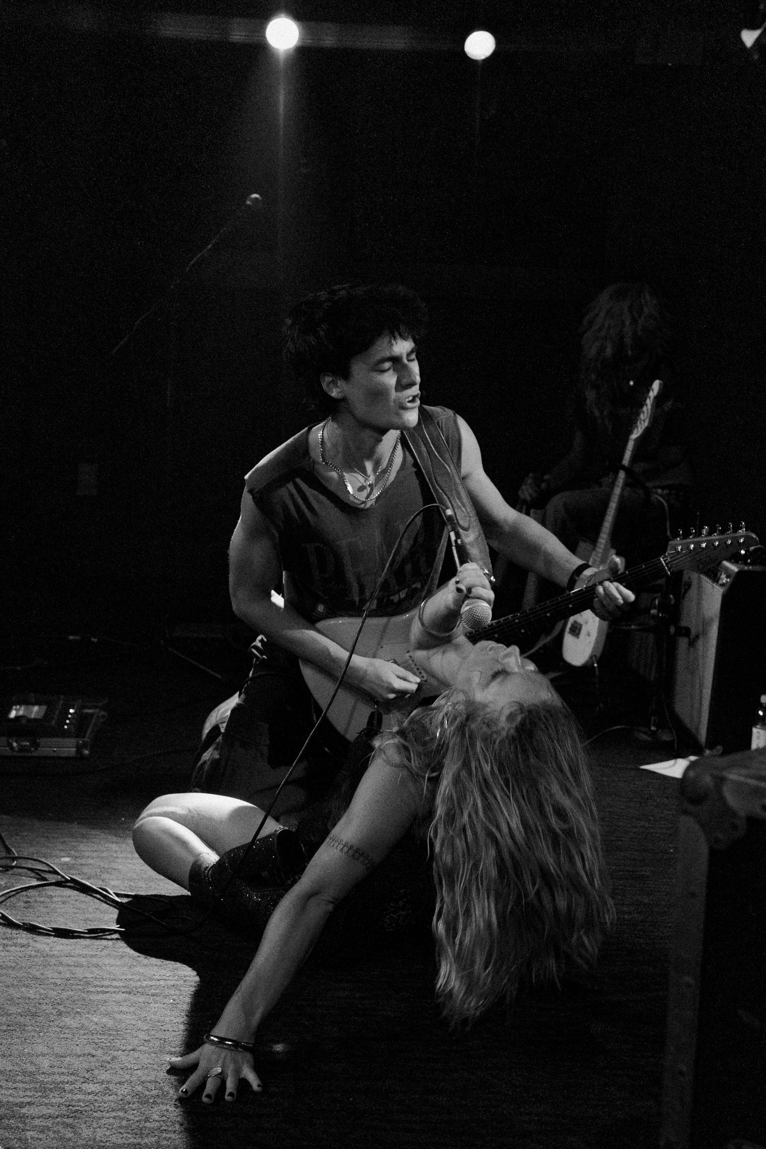 Black and white photo of two musicians performing on stage, one playing guitar while another sings dramatically on the floor.