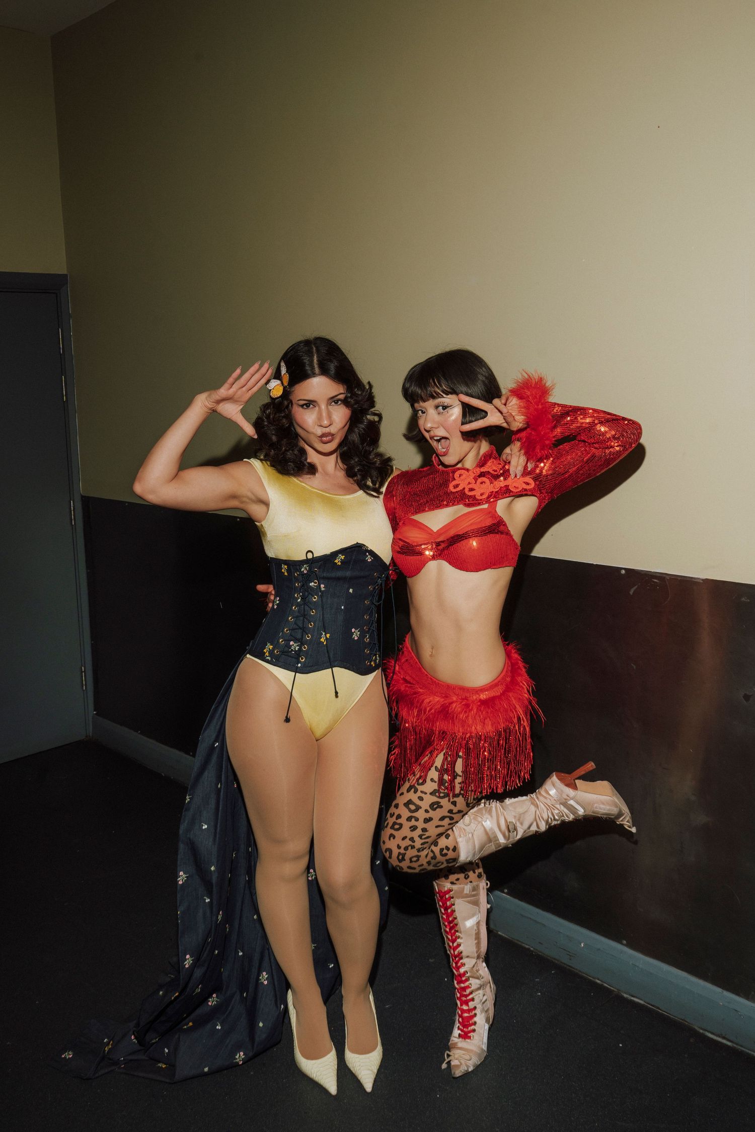 Two women in colorful burlesque costumes pose playfully in a hallway, one in yellow and blue, one in red and leopard print.