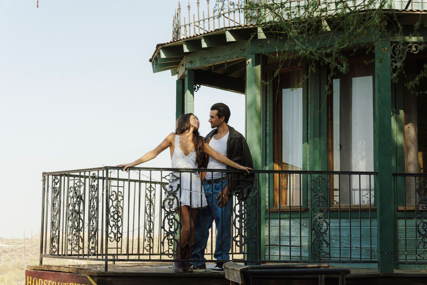 Romantic couple standing together on a vintage green balcony with ornate iron railings on a sunny day.