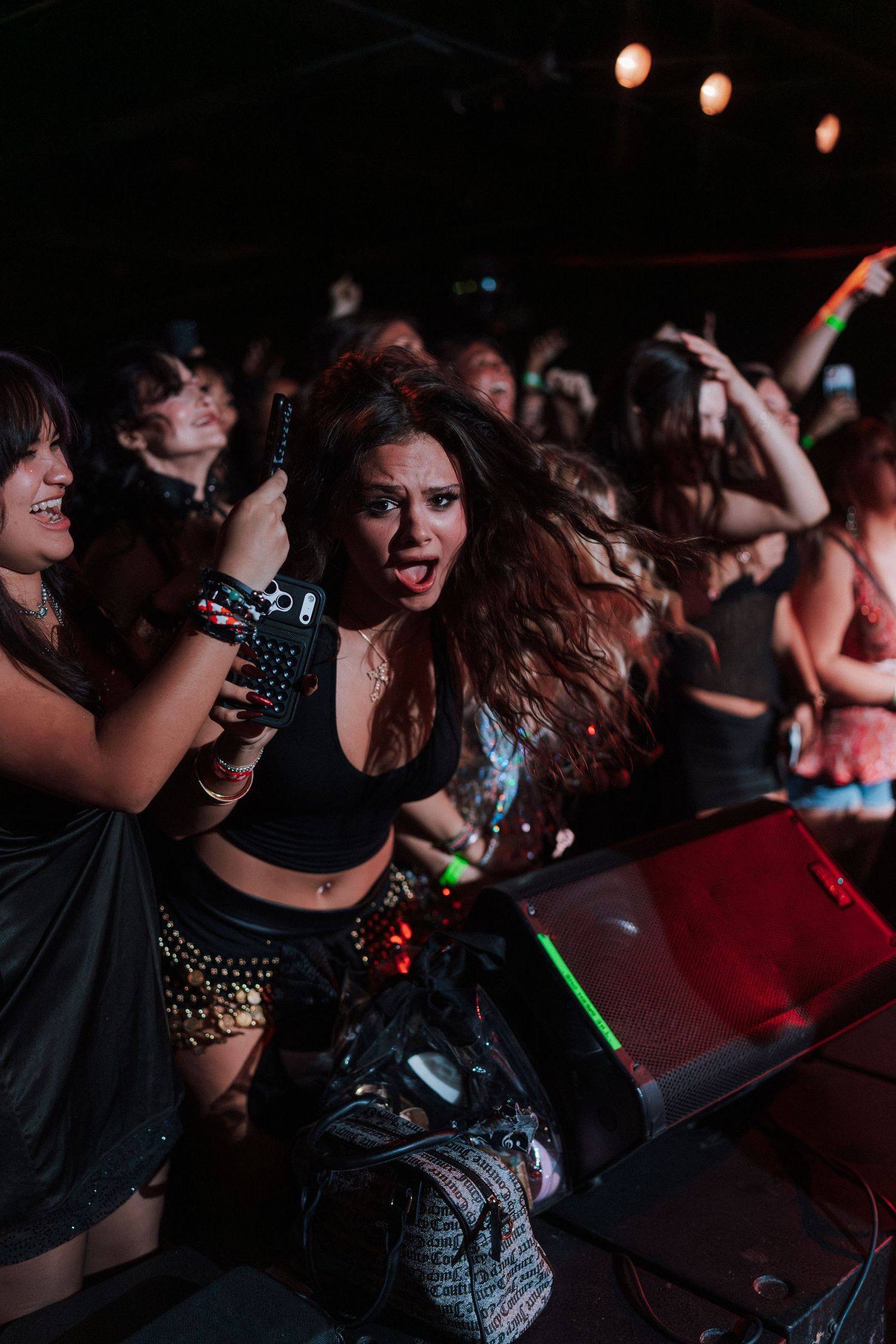 Excited young woman with flowing hair holding phone at a lively concert crowd with colorful lights.