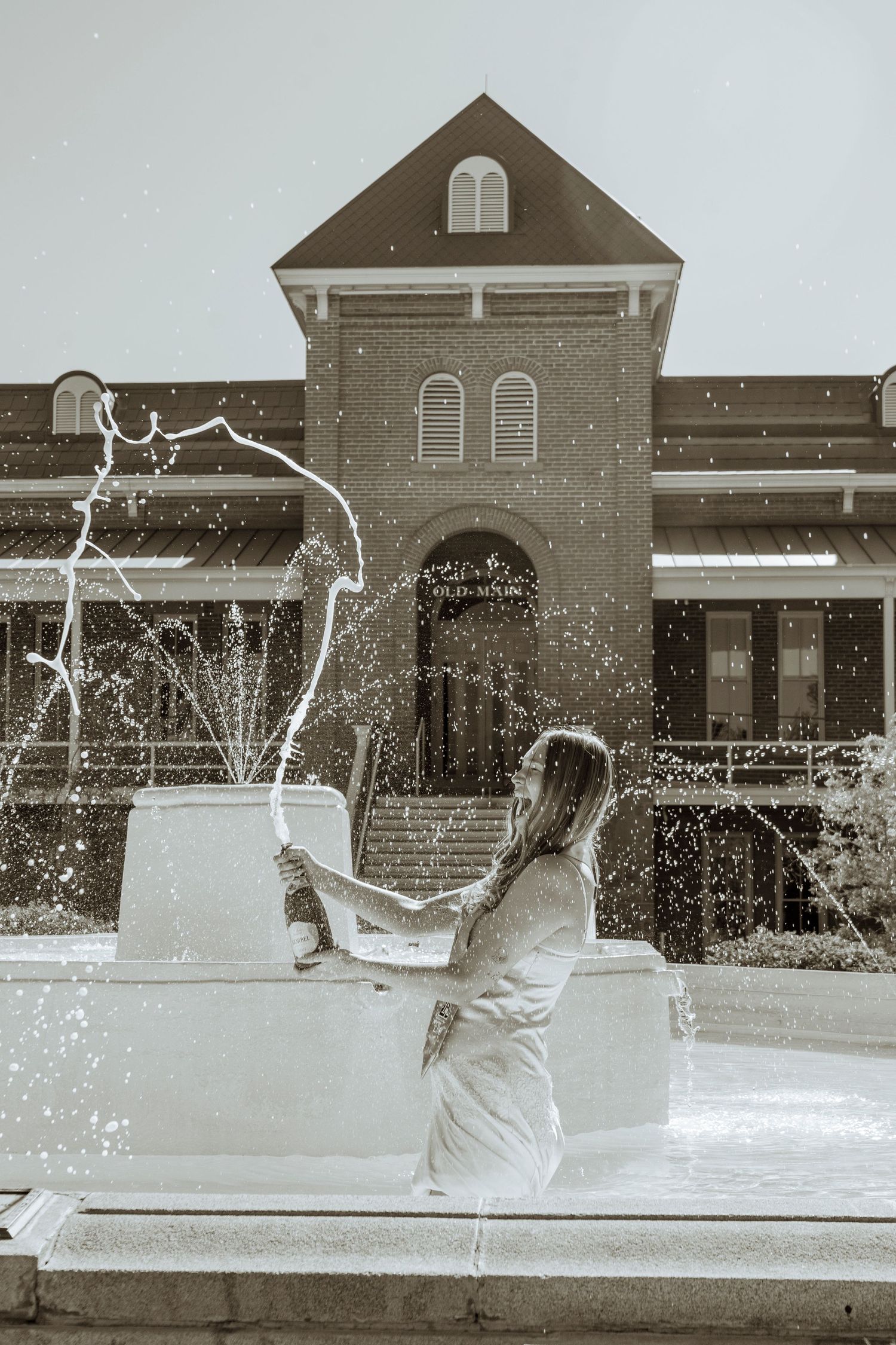Woman in white dress splashing in outdoor fountain near brick building in black and white photo.