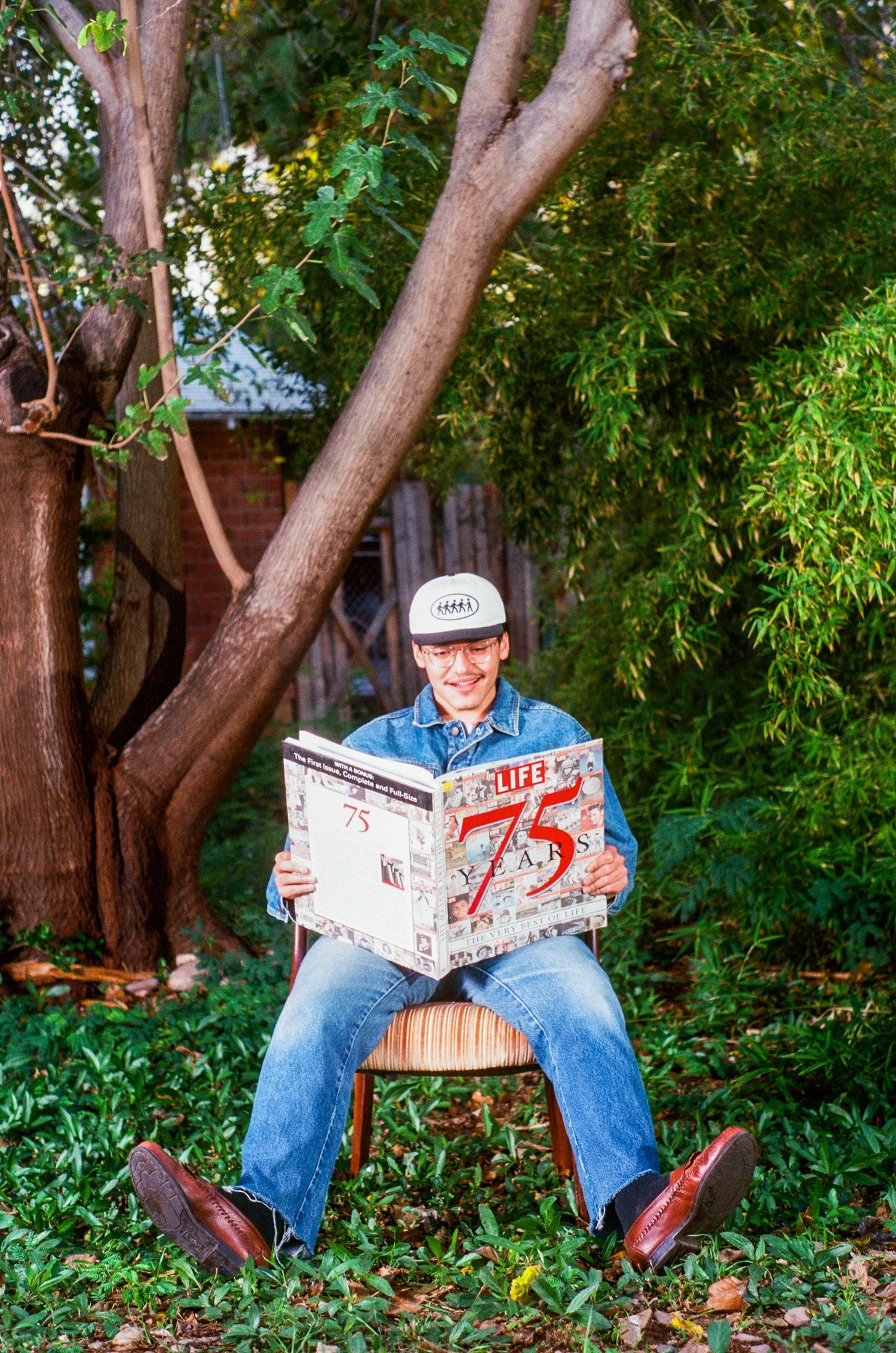Smiling man in denim outfit reads Life magazine issue 75 while seated outdoors.