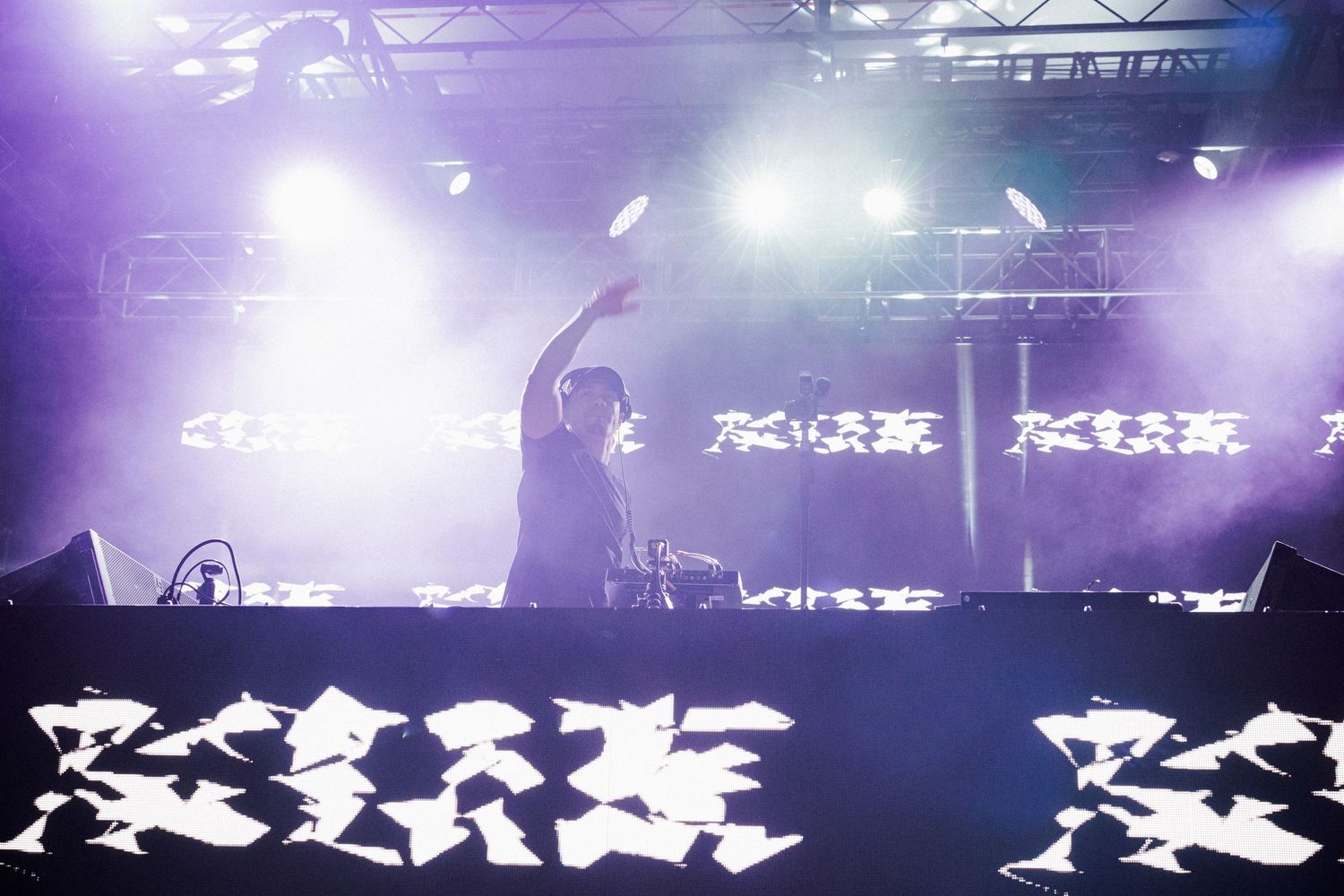 DJ performing at illuminated booth on stage with purple fog and bright graphic backdrop.