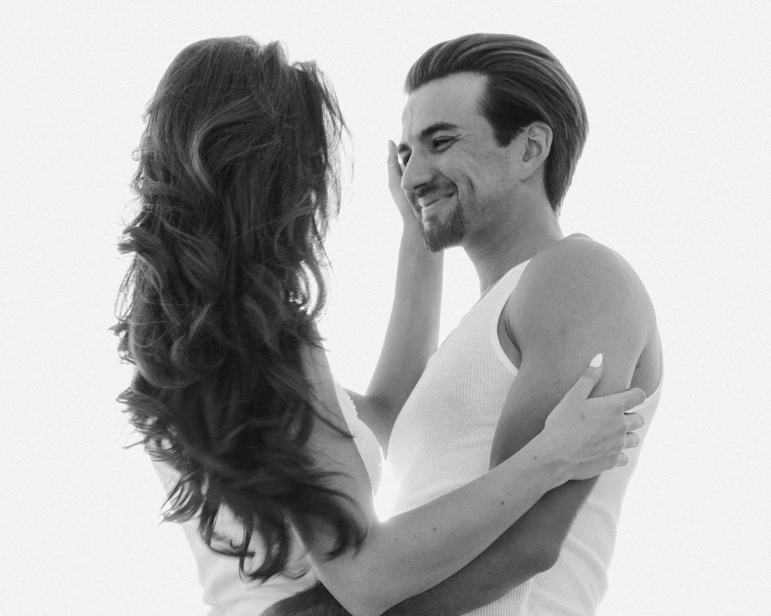 Black and white photo of smiling man embracing woman with long dark hair on white background.