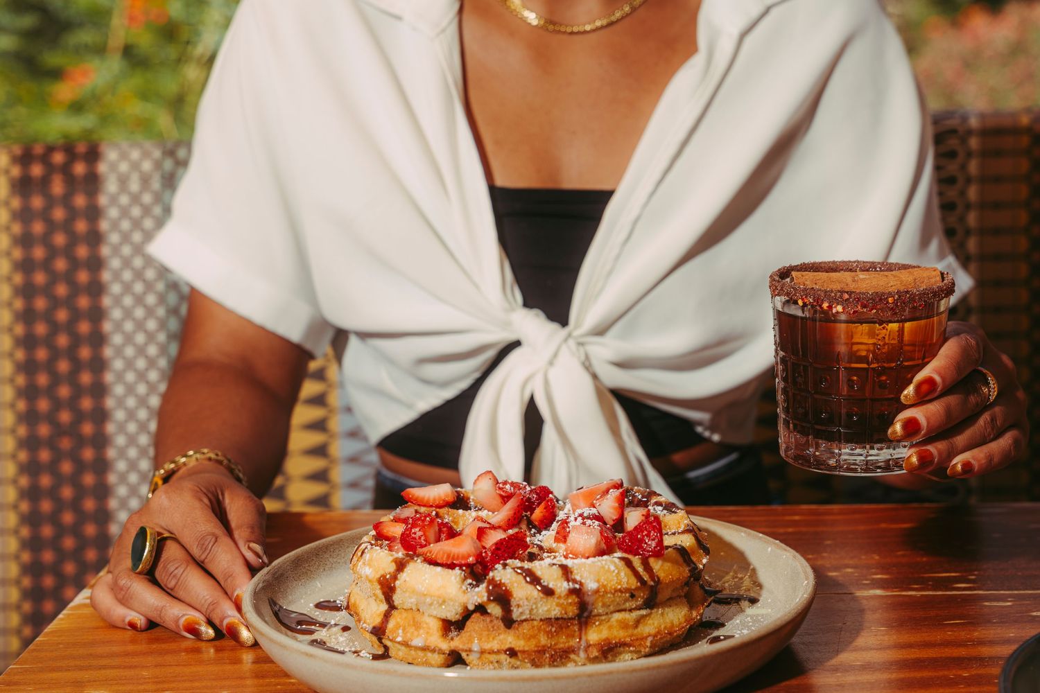 Woman holding a stack of strawberry waffles and a cocktail at an outdoor restaurant.