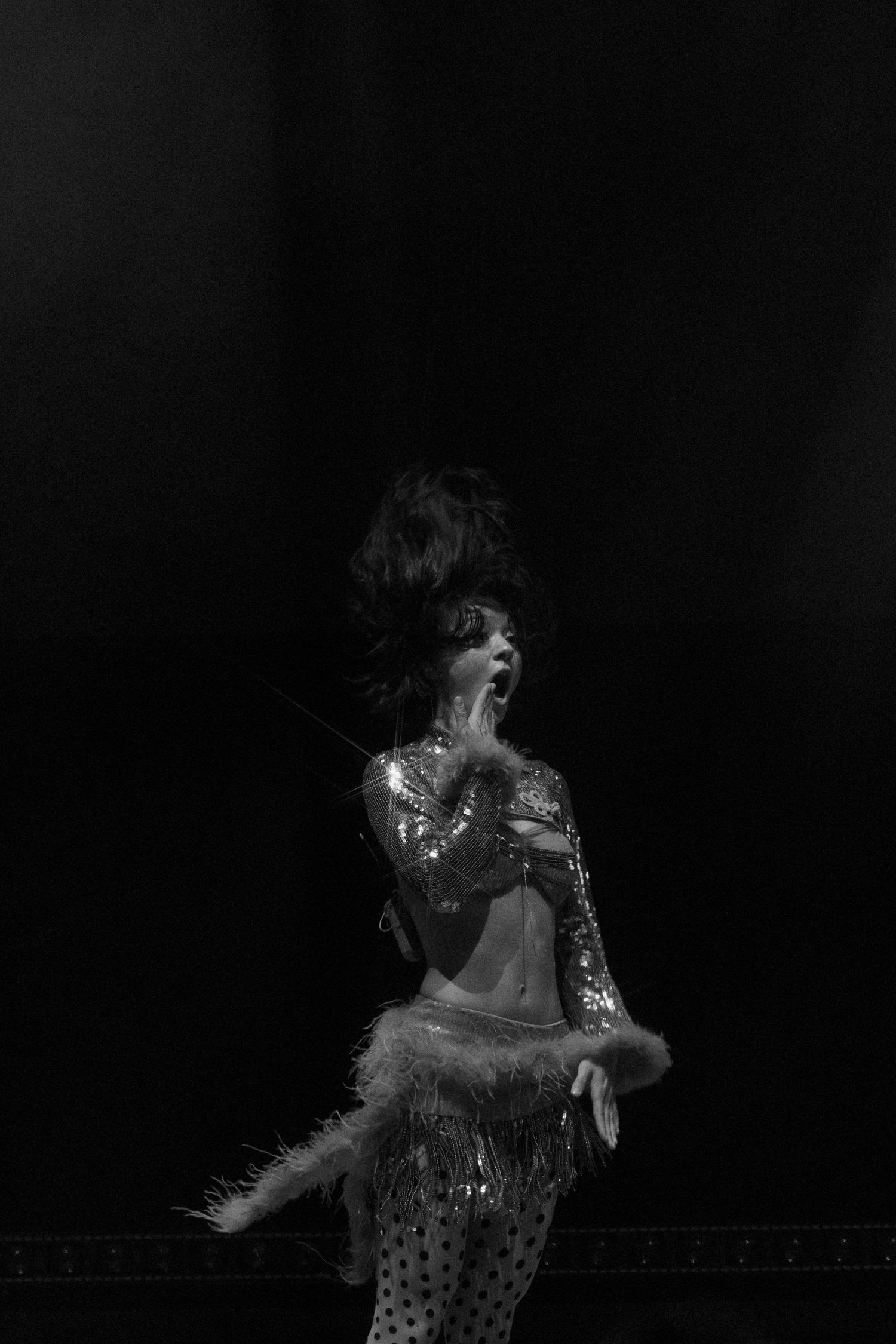 Black and white photo of a glamorous female performer on stage in sequined costume with feathers and dramatic pose.