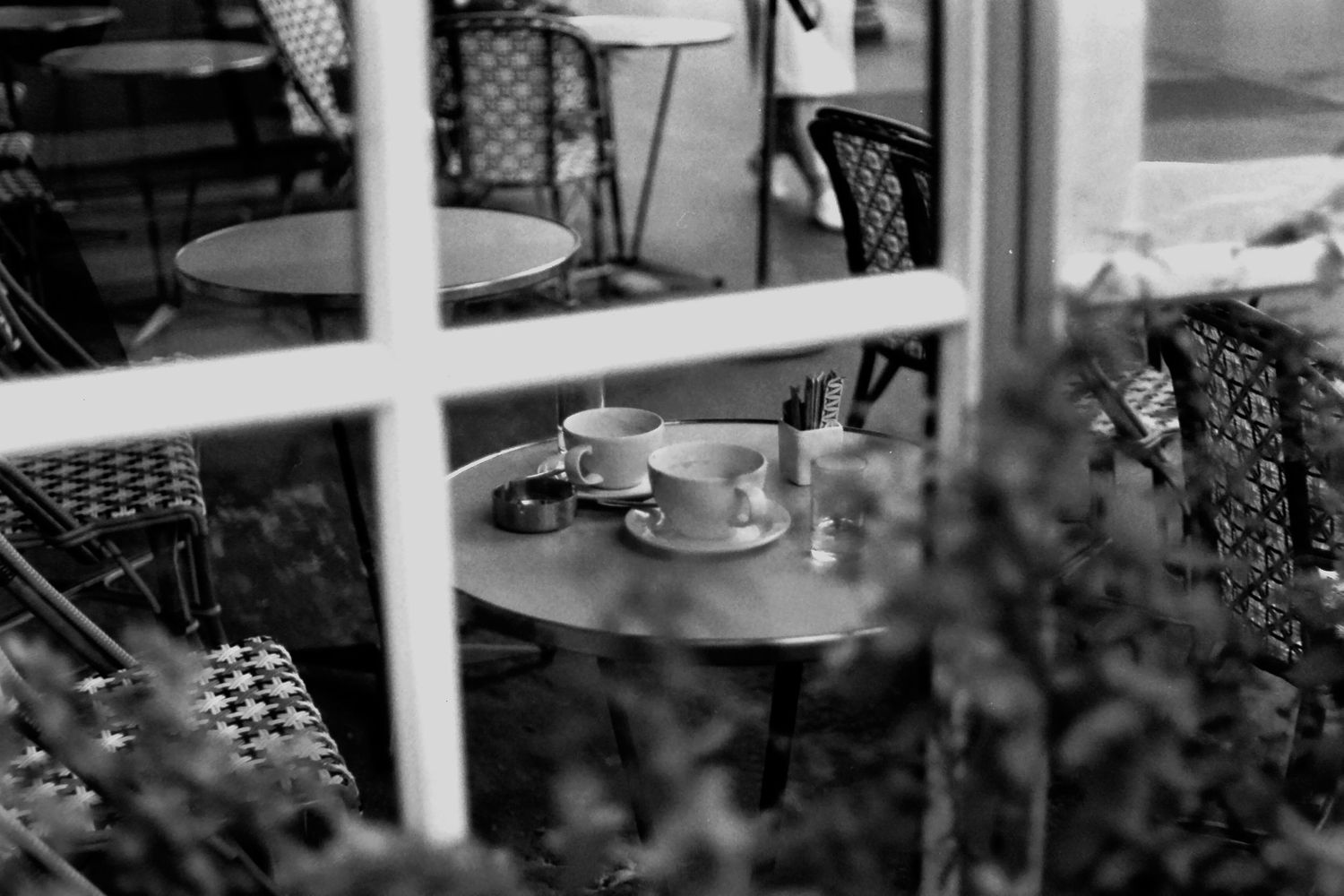 Black and white photo of a cozy cafe patio table with two coffee cups viewed through a window.