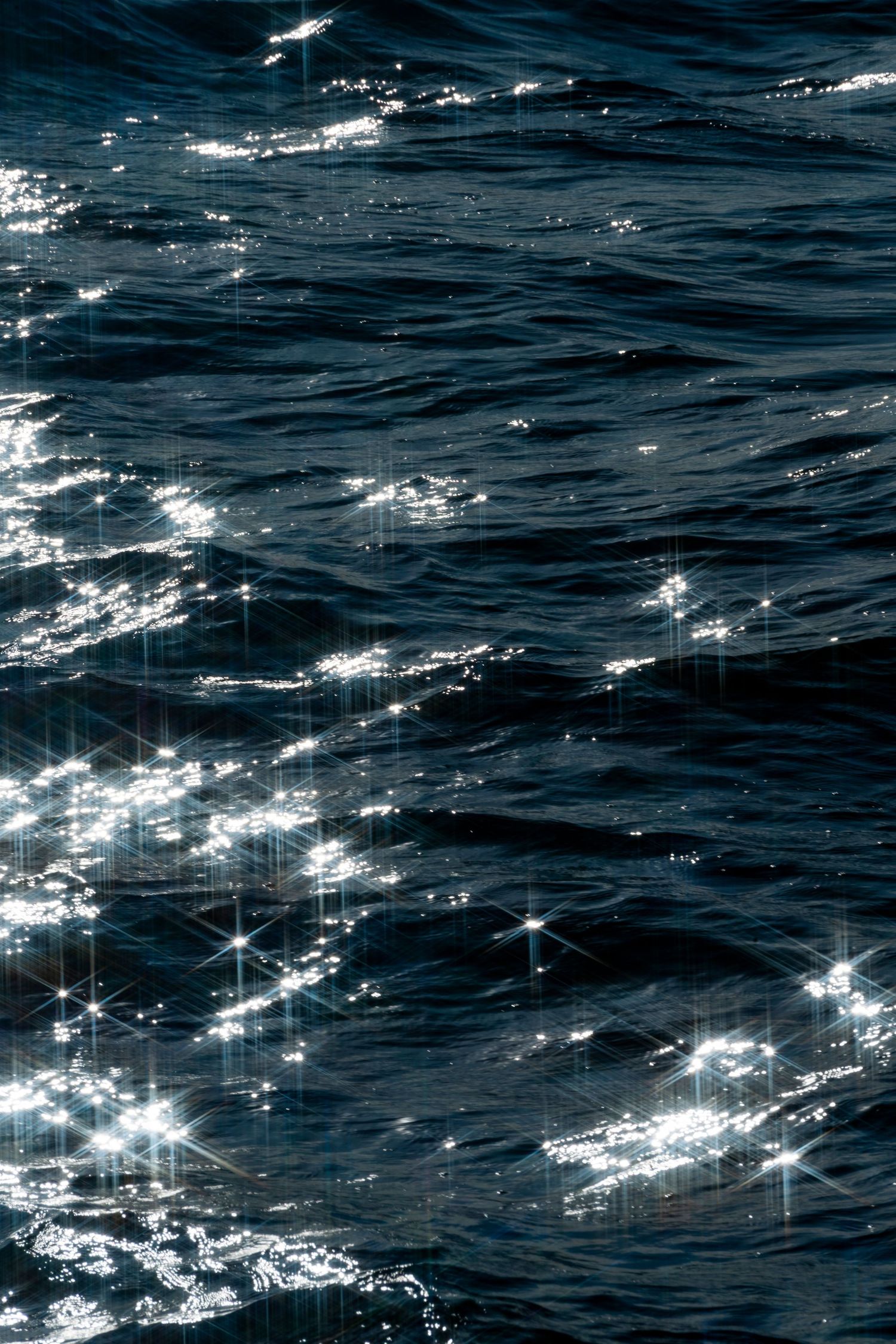Sunlight sparkles on dark blue ocean waves, creating dazzling star-shaped reflections on the rippling water surface.