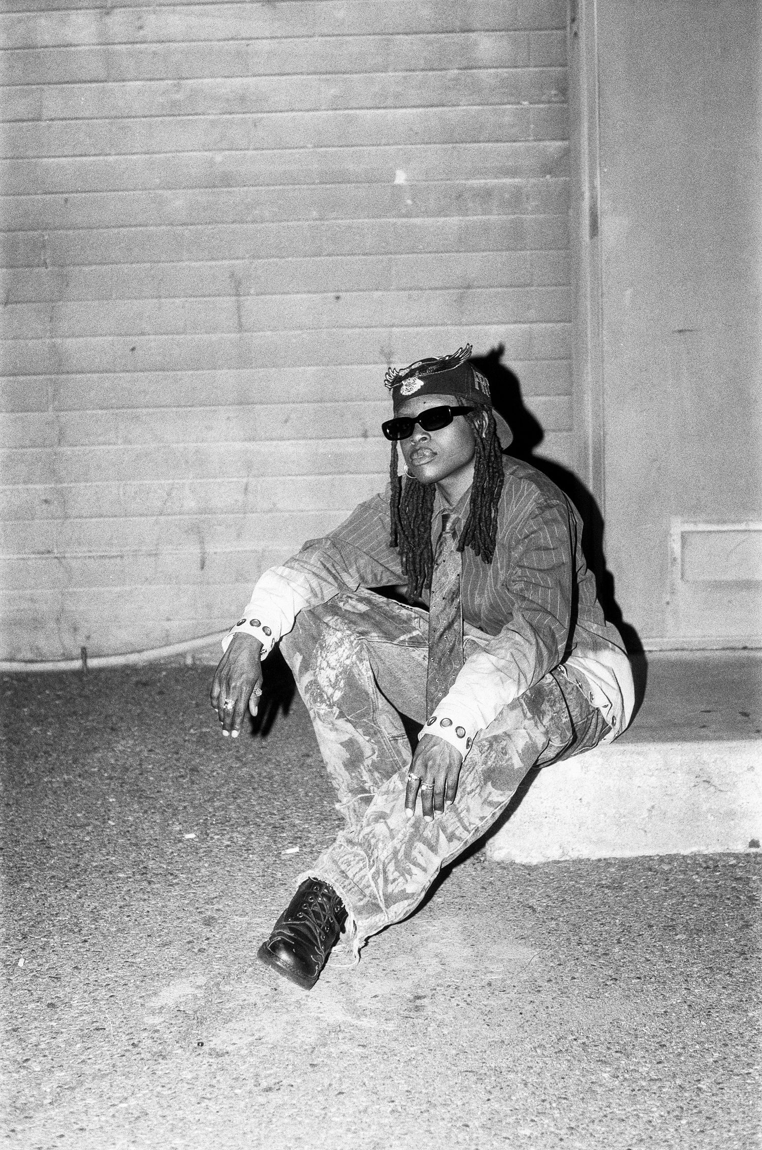 Black and white photo of person with braids and sunglasses sitting on ground outdoors.