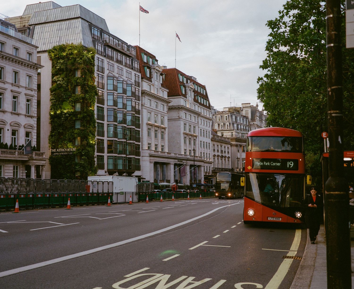 Red double-decker bus on a quiet London street lined with classic architecture and trees on a cloudy day.