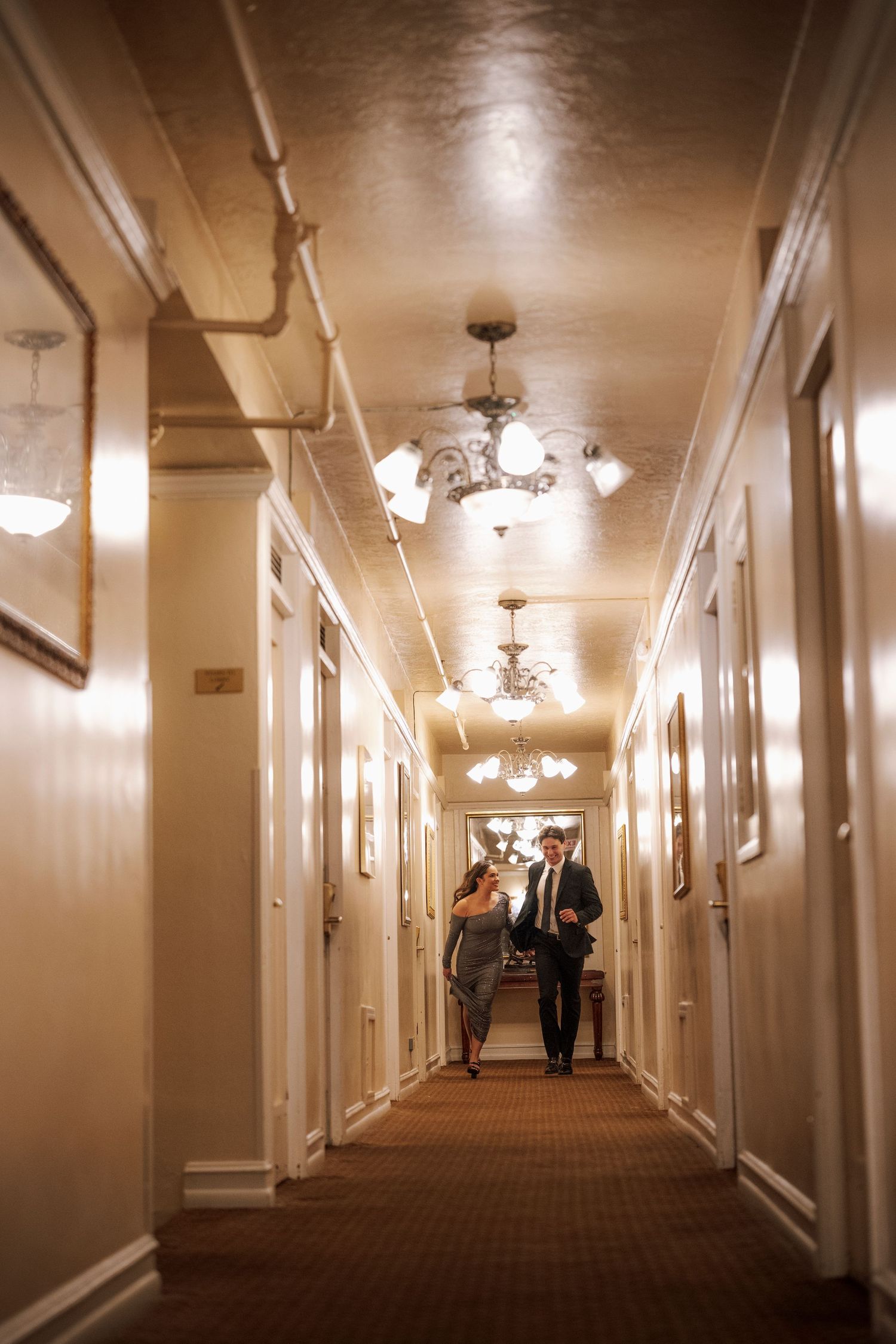 Couple walking together down a warmly lit vintage hotel hallway with chandeliers.