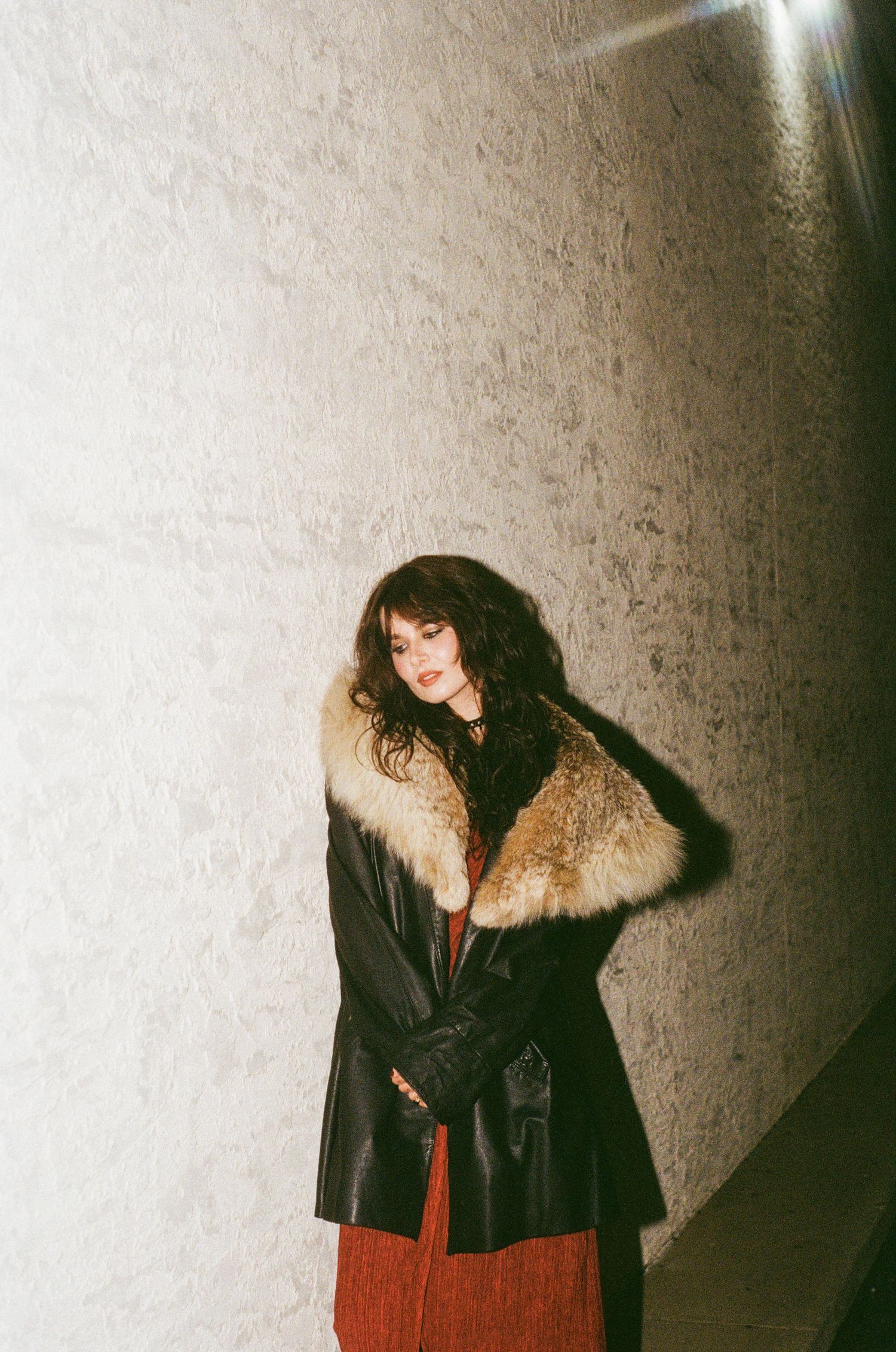 Woman in black leather coat with fur collar posing against a textured wall in dramatic lighting.