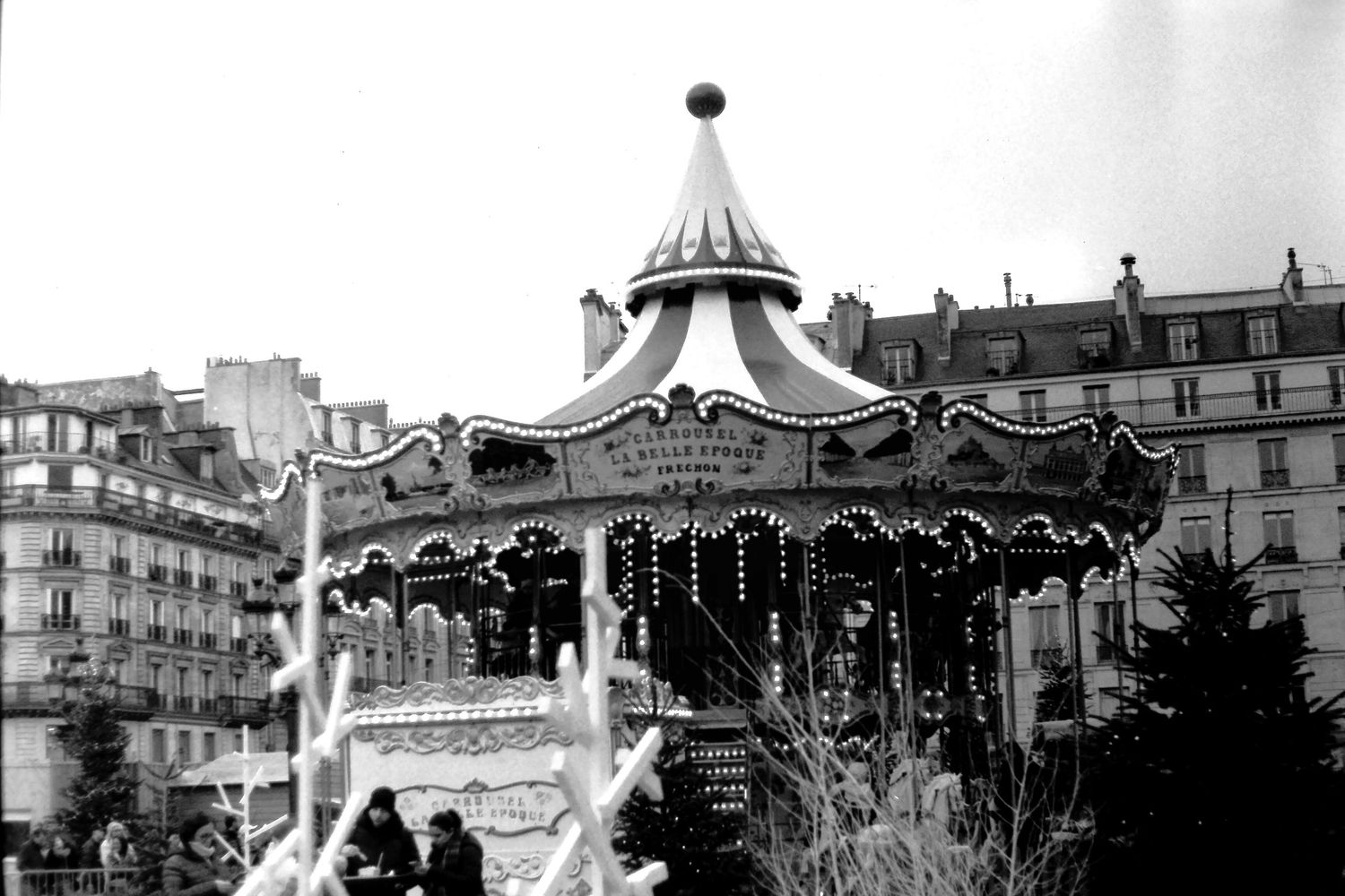 Vintage black and white photo of a classic carousel in Paris, France, with ornate details and city buildings in background.