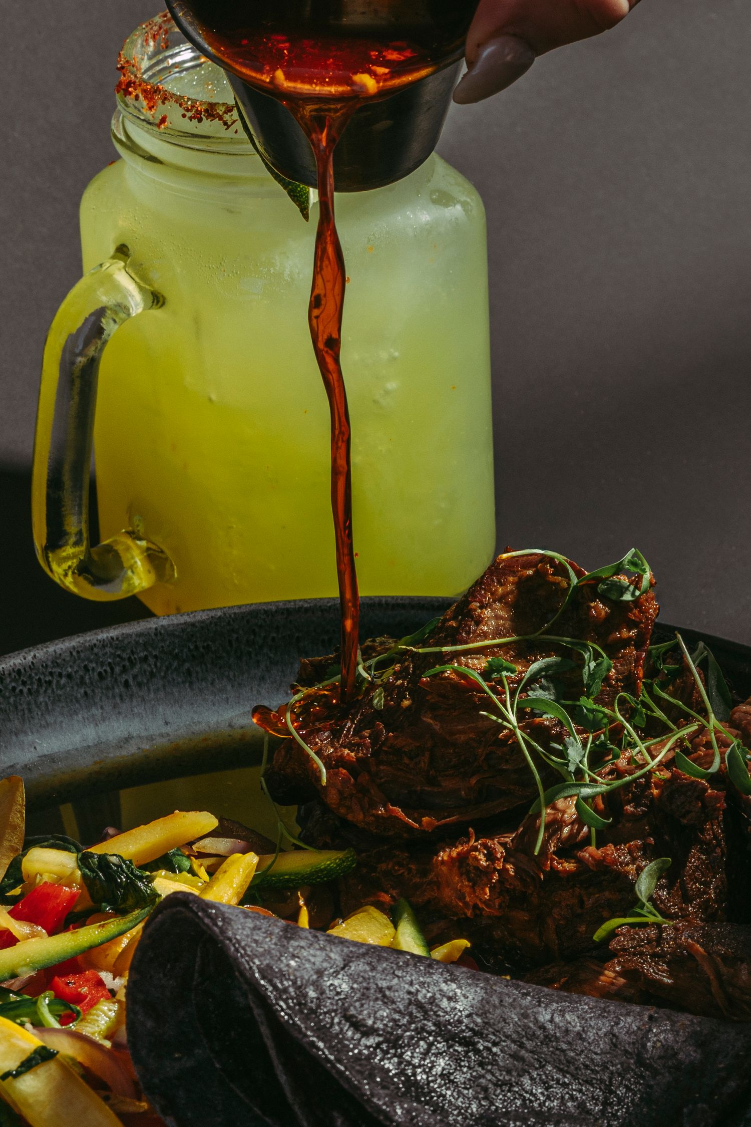 Dark red sauce being poured over a spiced meat dish with fries and greens.