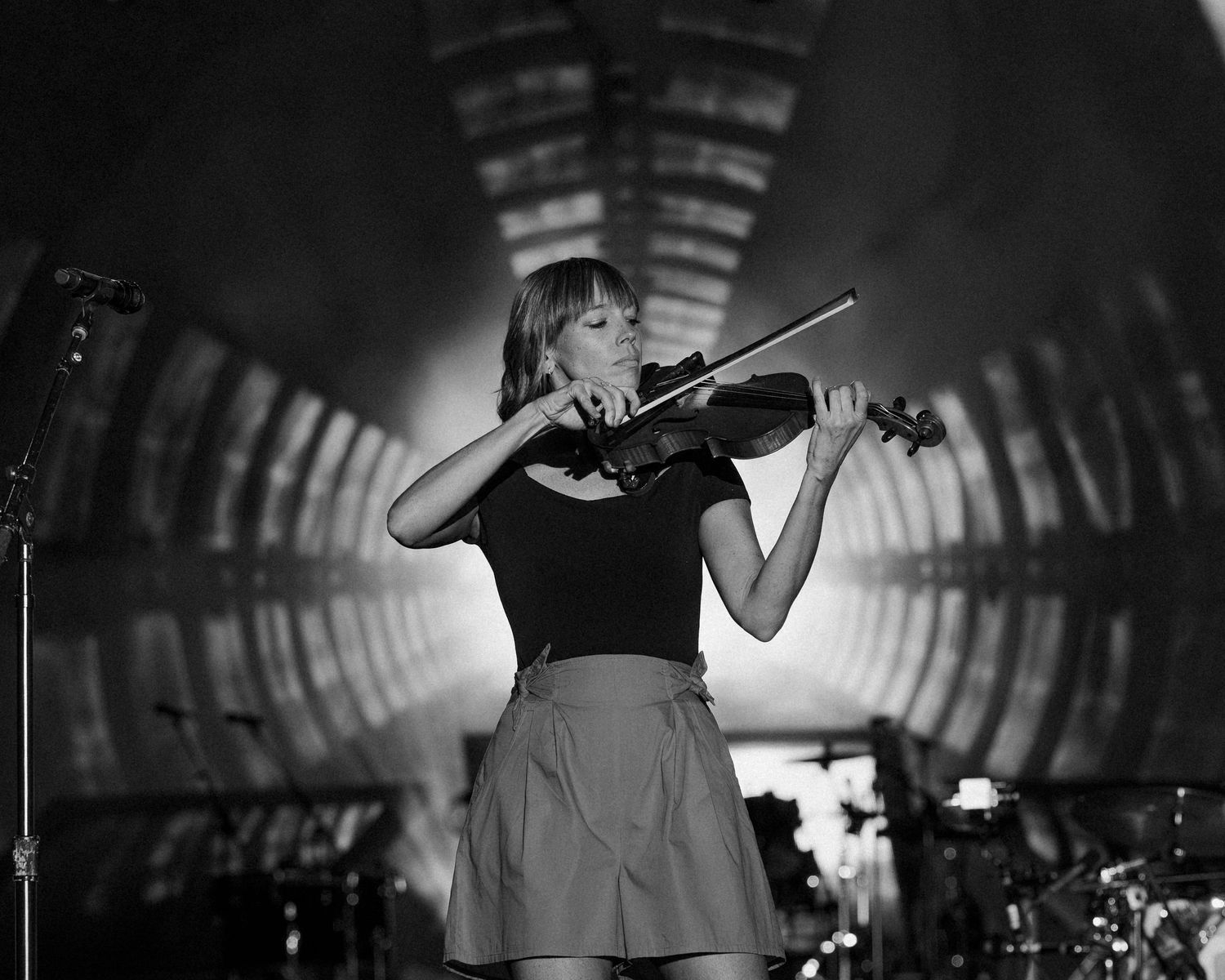 Black and white photo of a female violinist performing on stage with dramatic tunnel lighting backdrop.