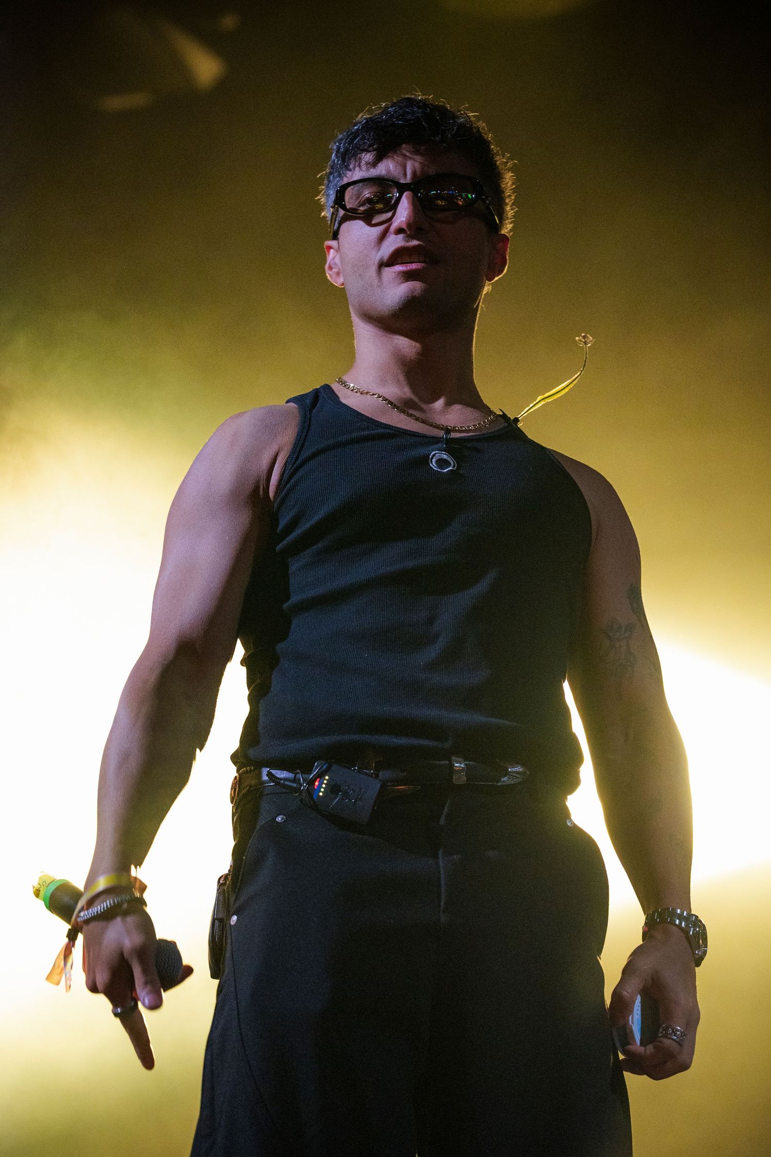 Male performer in black sleeveless top and sunglasses standing on stage in golden spotlight.