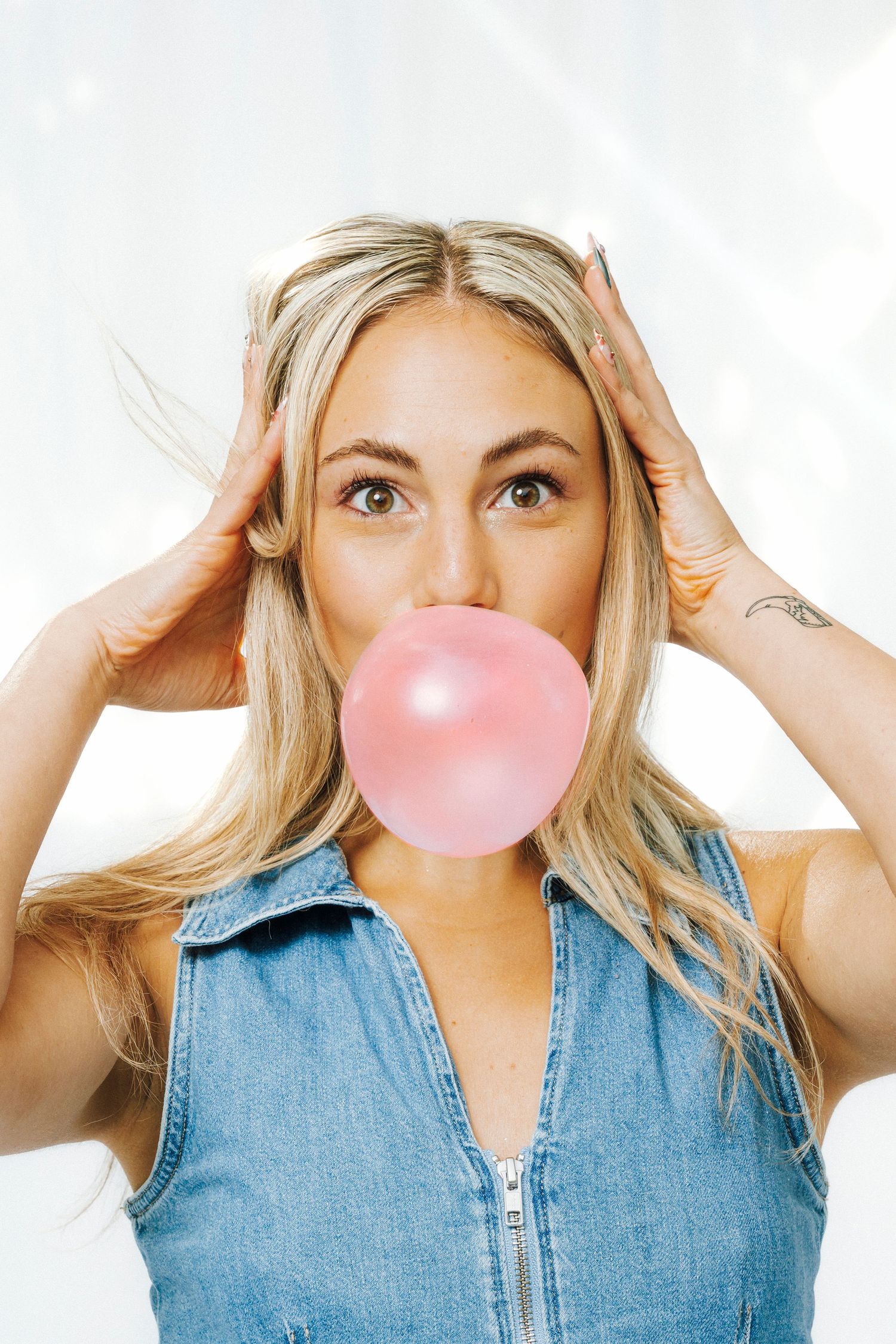 Blonde woman in denim vest blowing large pink bubble gum bubble with hands raised to head.